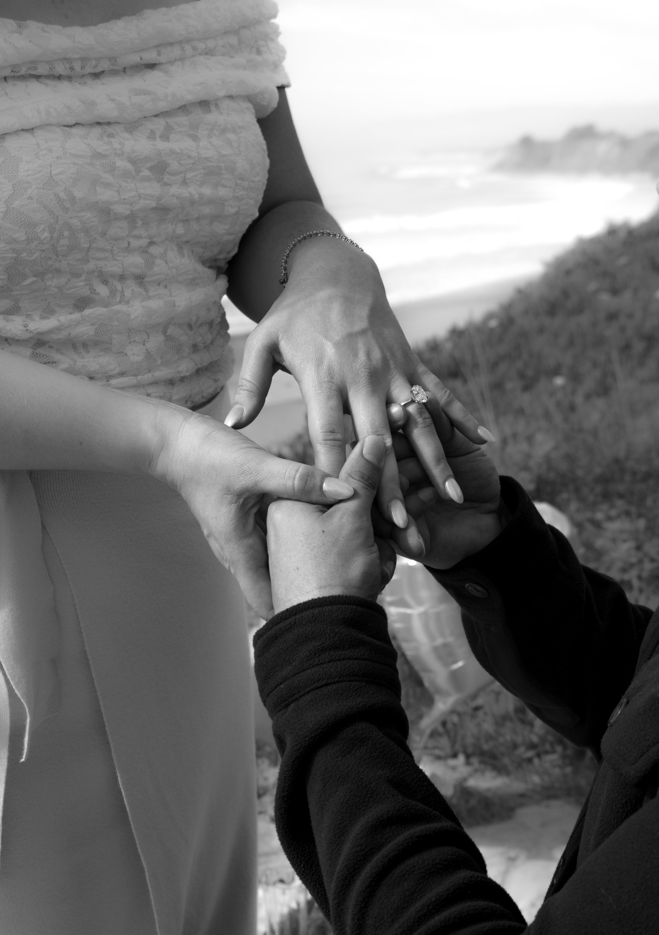 A black-and-white photo of a man proposing marriage to a woman by placing a ring on her finger. The woman's hand is adorned with a ring and a bracelet, and there is a scenic background of the beach in Santa Barbara.