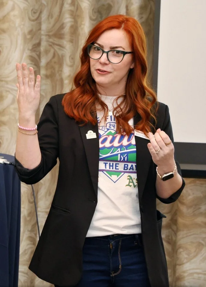 A woman with red hair and glasses wearing a black blazer over a white t-shirt with a colorful logo. She is speaking or presenting, using hand gestures, in front of a beige marble wall and a presentation screen.