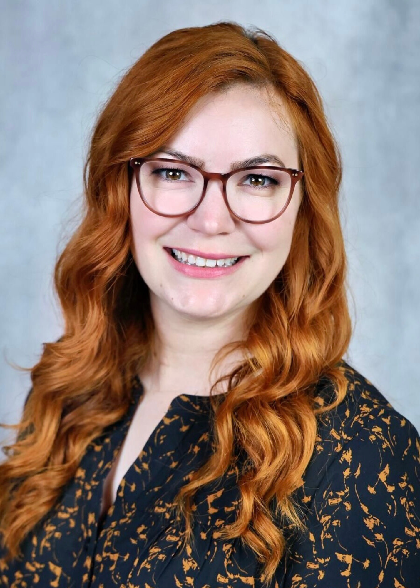 A young woman with long, wavy red hair, wearing large brown glasses and a dark blouse with an orange floral pattern, smiling at the camera against a plain background.