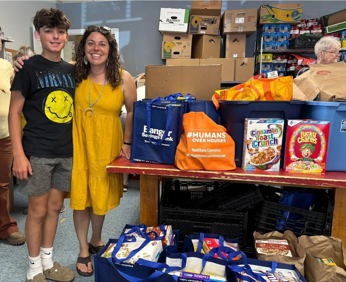 A young man and a woman standing together at a food bank with boxes and bags of food and supplies on a table and shelves behind them.