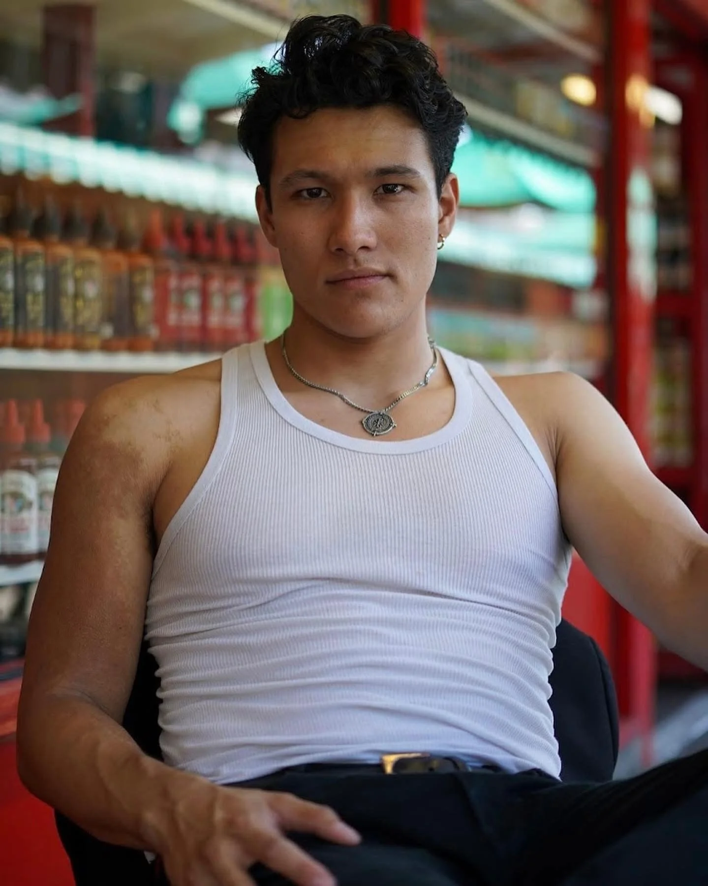 Young man with dark, curly hair, wearing a white tank top and silver jewelry, sitting indoors near shelves with bottled products, with a serious expression.