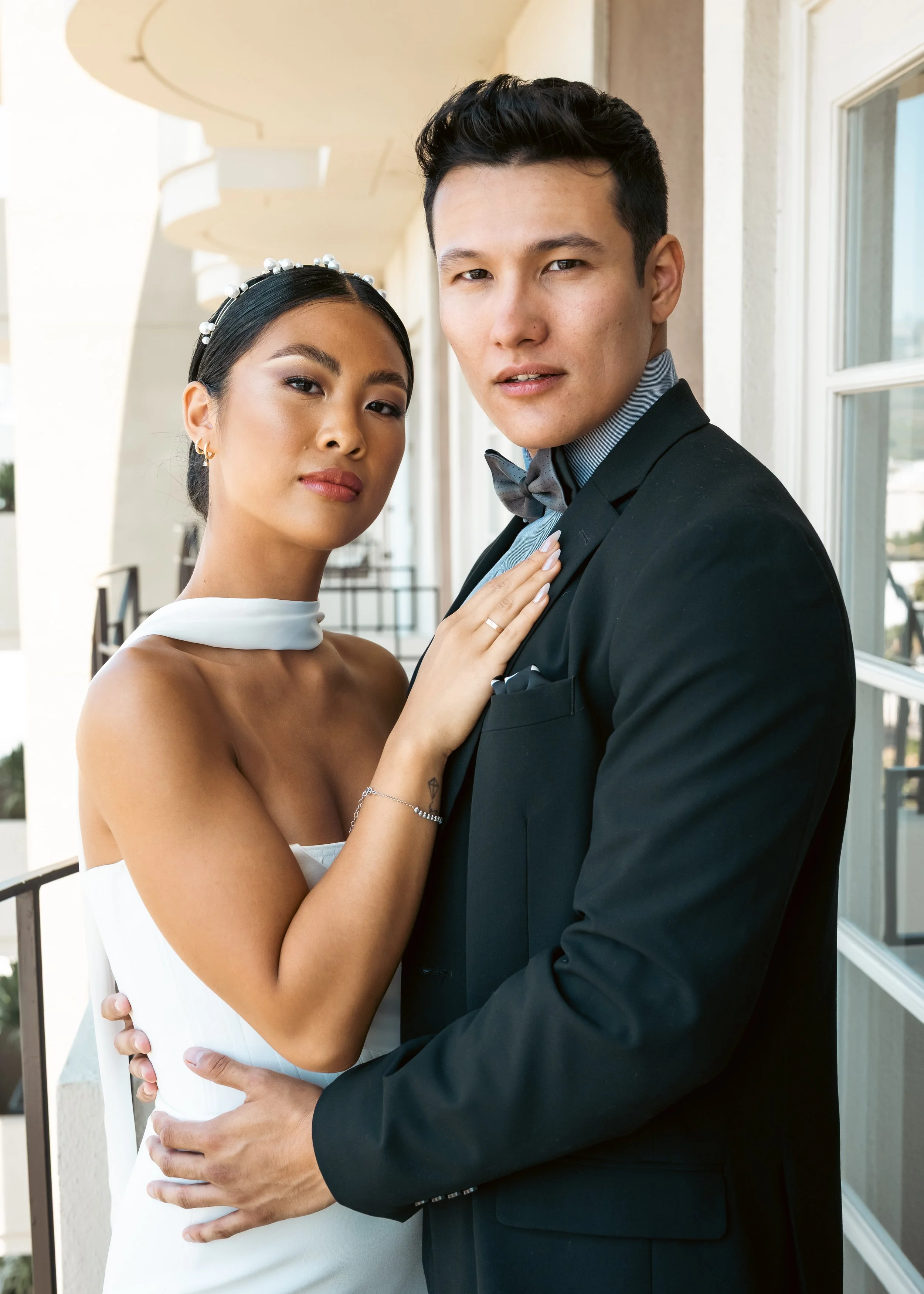 A couple dressed in formal wedding attire, standing on a balcony with white walls and large windows, looking into the camera.