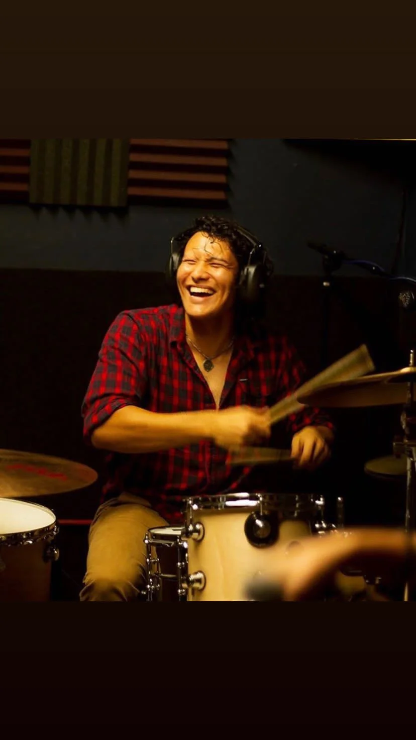 A person playing drums, smiling and laughing, wearing a red and black plaid shirt and headphones in a music studio.