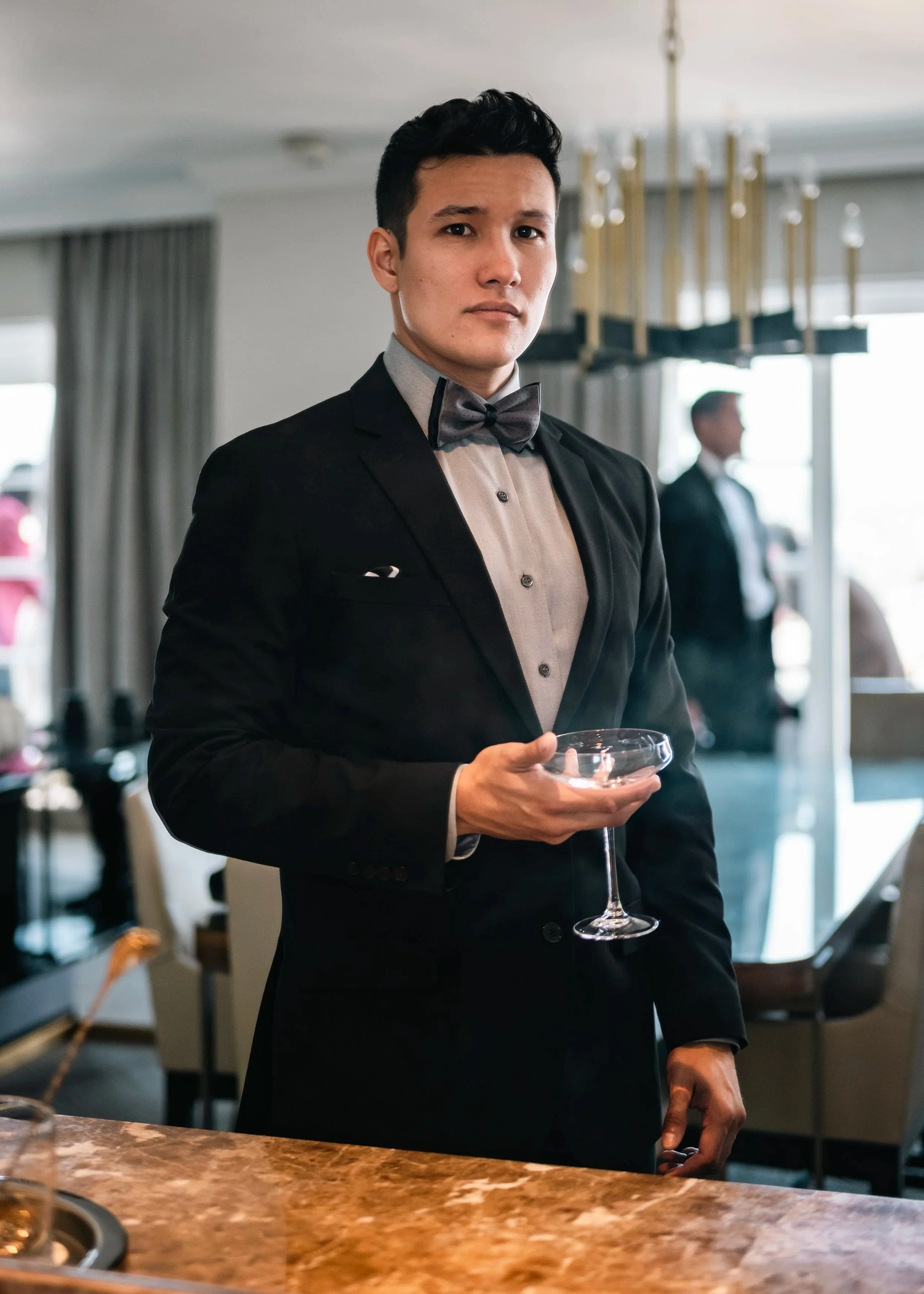 A young man in a tuxedo with a bow tie holding a coupe glass, standing in an upscale restaurant or event setting.