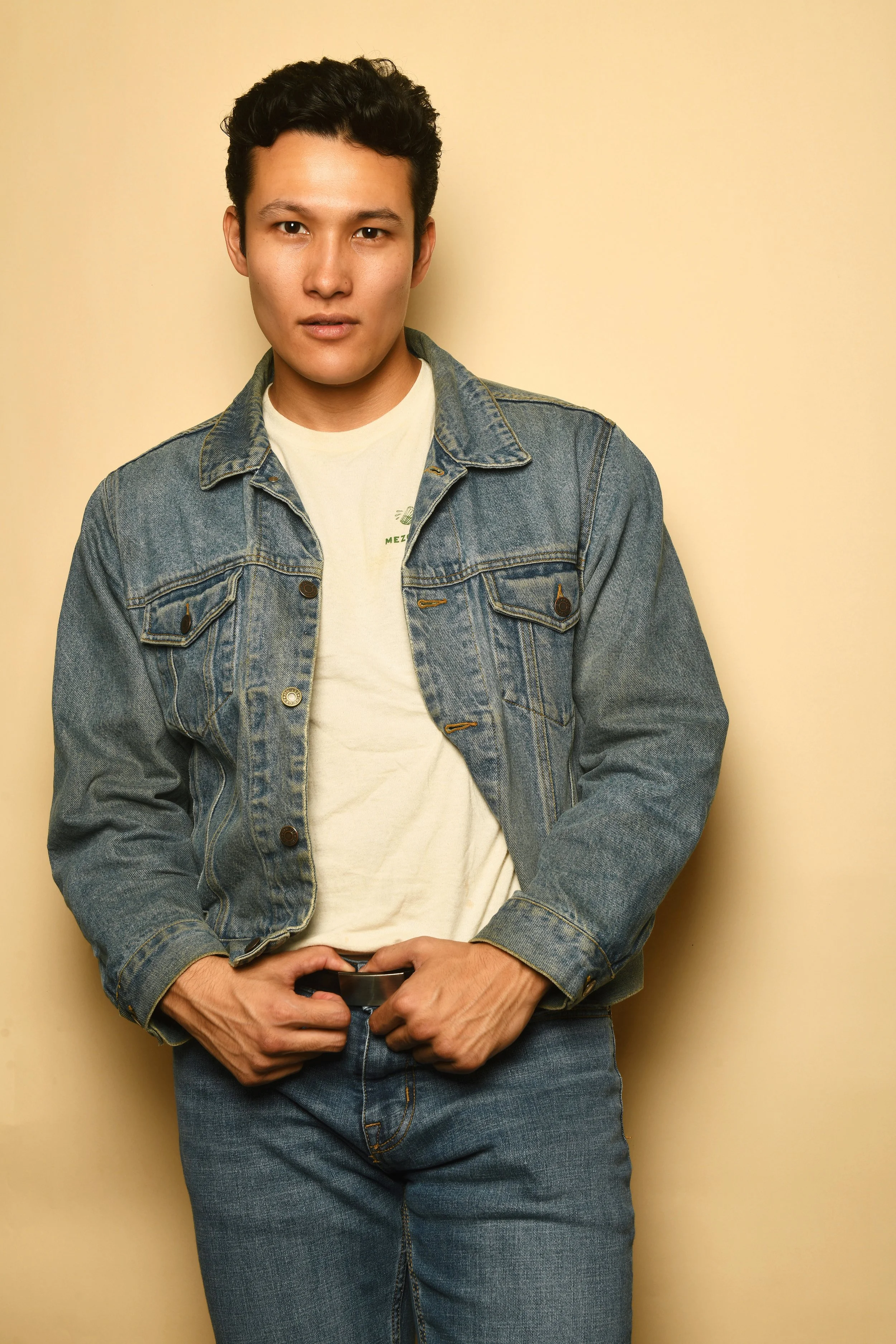 A young man with short black hair standing against a plain yellow wall, wearing a denim jacket over a white T-shirt and blue jeans, looking directly at the camera with a neutral expression.