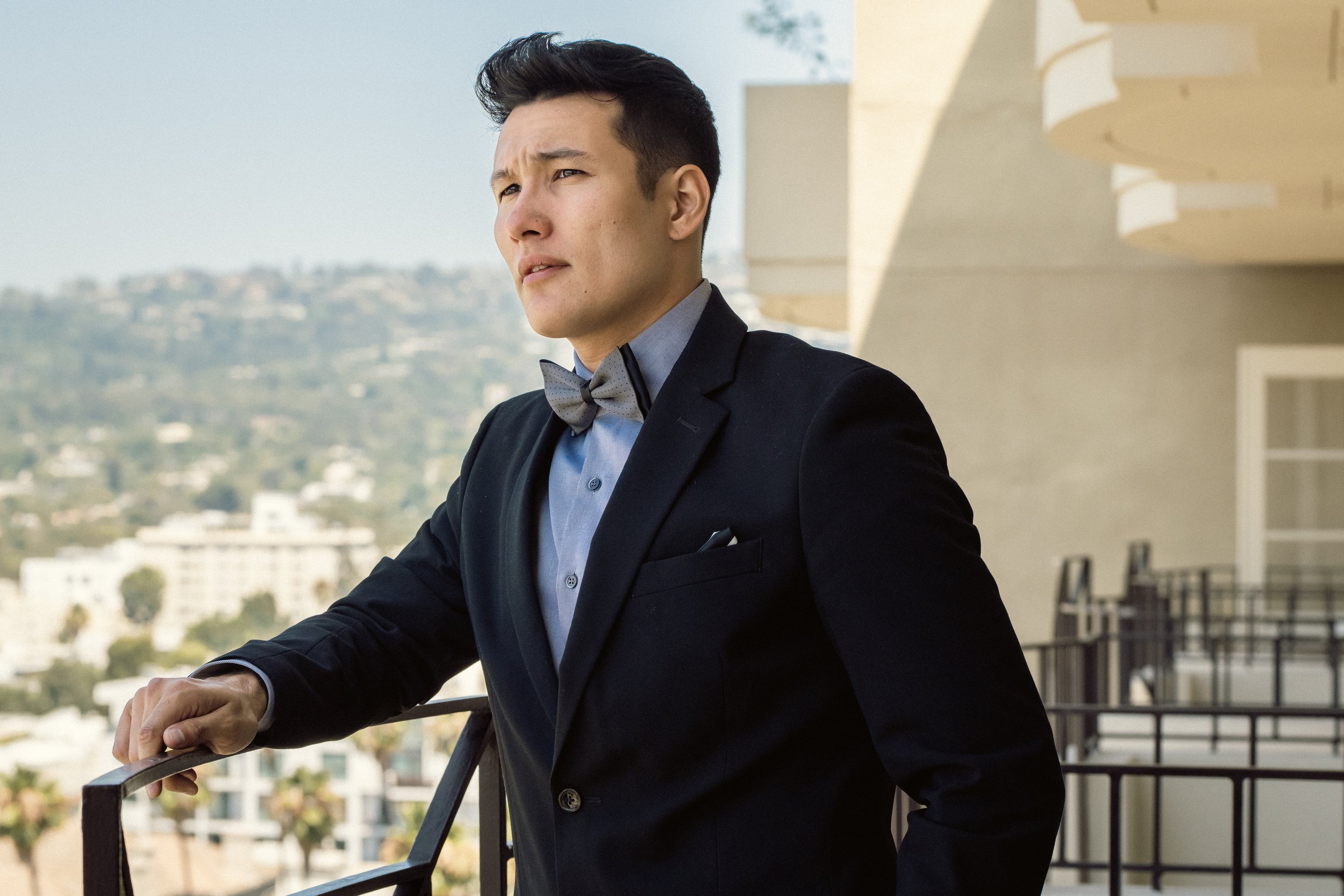 A young man in a tuxedo with a bow tie, looking into the distance on a balcony with a cityscape in the background.