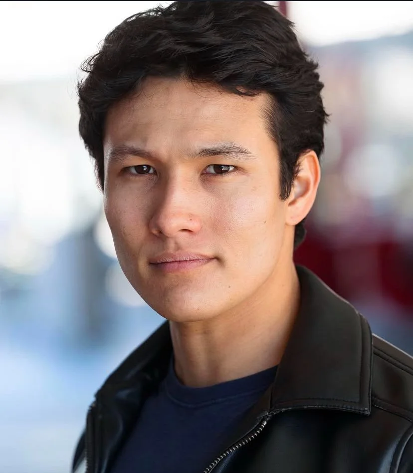 A young man with dark hair wearing a black leather jacket, looking at the camera, outdoors in a city setting.