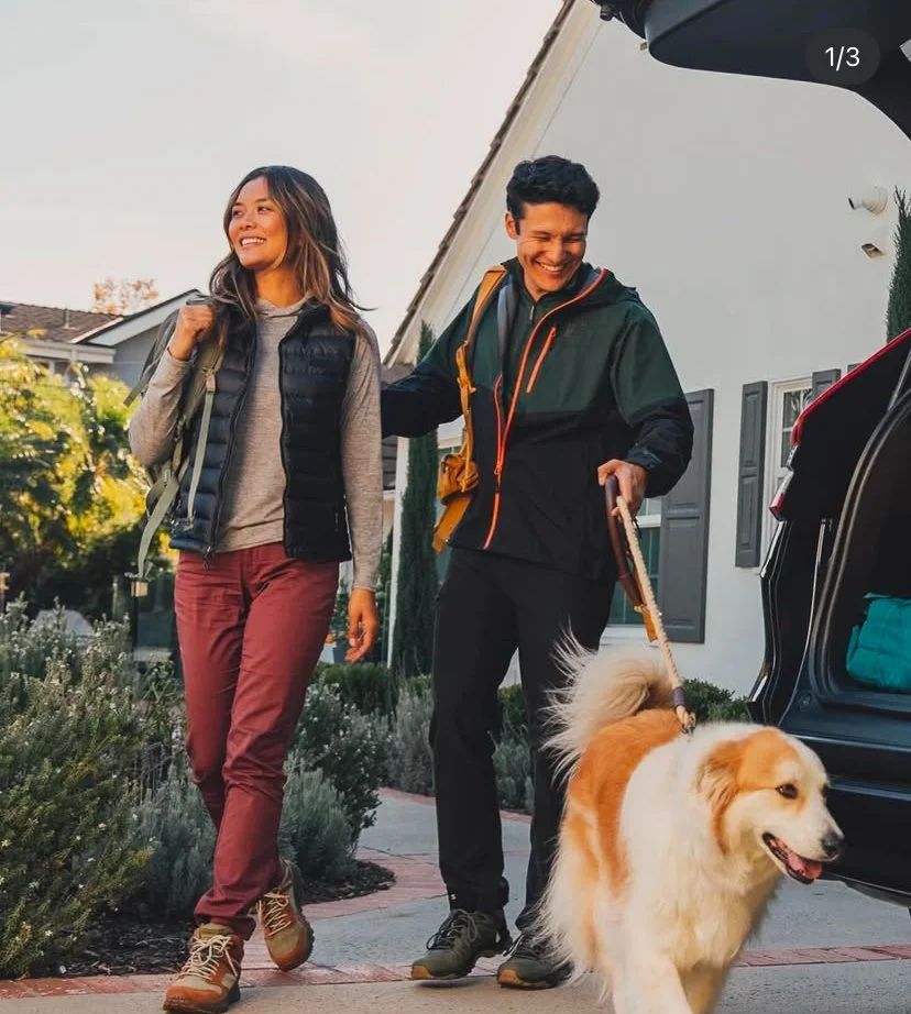 A woman and a man smiling and walking with a dog near a house. The woman is wearing a gray sweater, a black vest, and red pants, while the man is in a black jacket and dark pants, holding a leash attached to a large, fluffy, cream-colored dog.