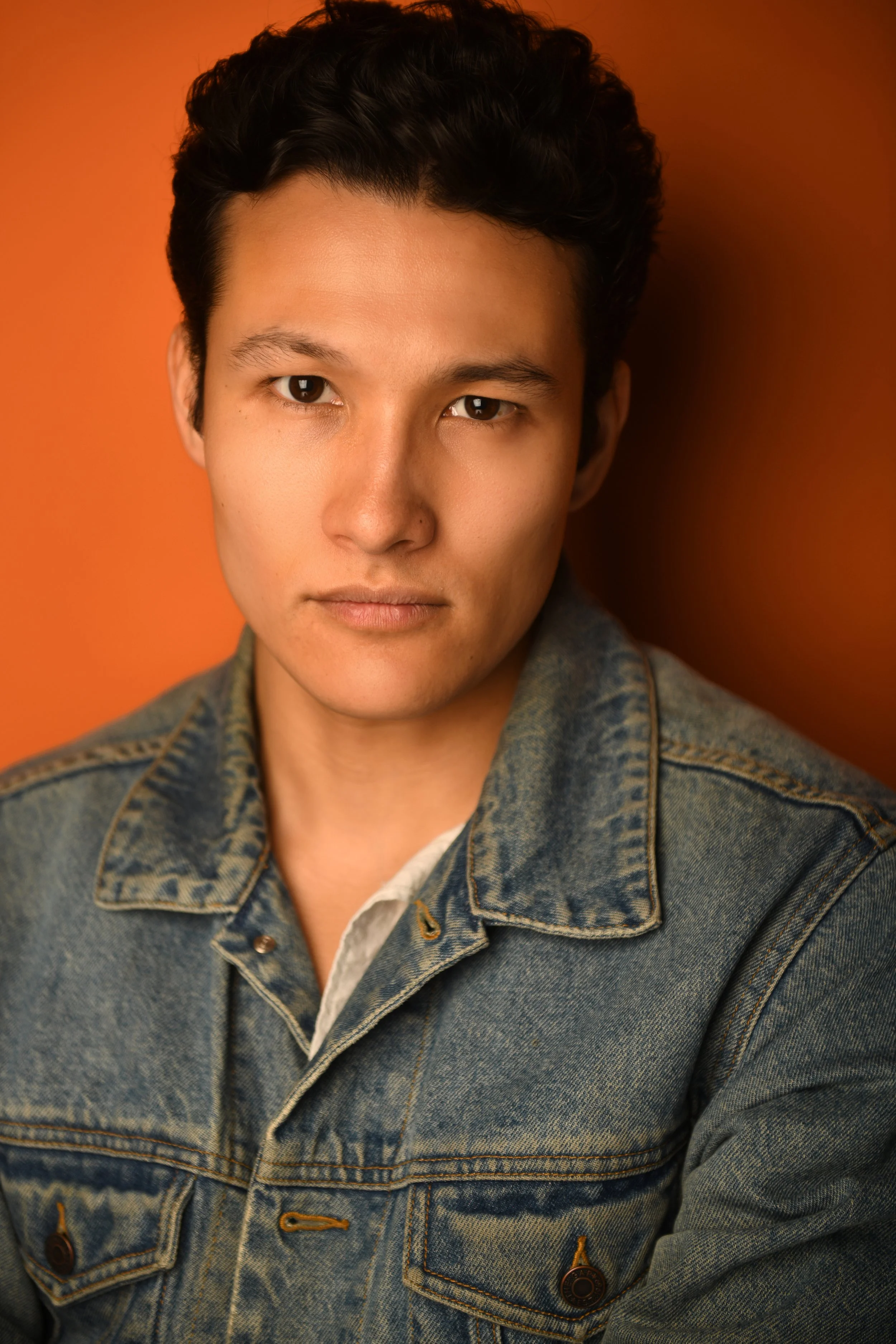 A young man with dark, curly hair wearing a denim jacket, looking directly at the camera with a serious expression, against an orange background.