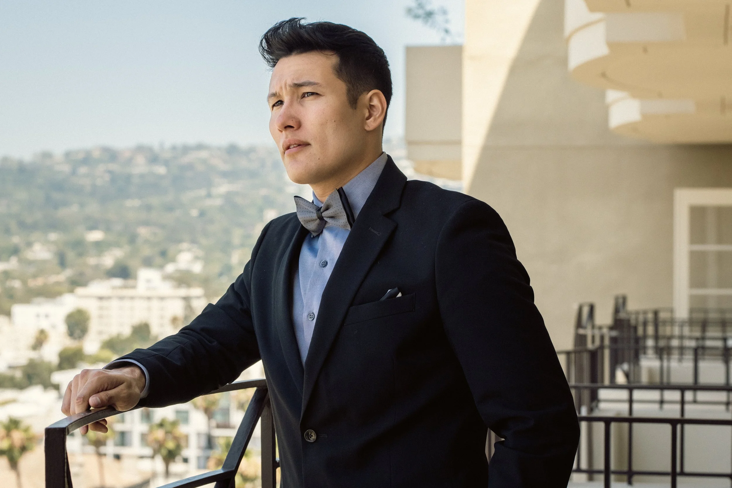 A young man dressed in a black suit, blue shirt, and gray bow tie, standing on a balcony with a cityscape and hillside in the background, gazing thoughtfully into the distance.