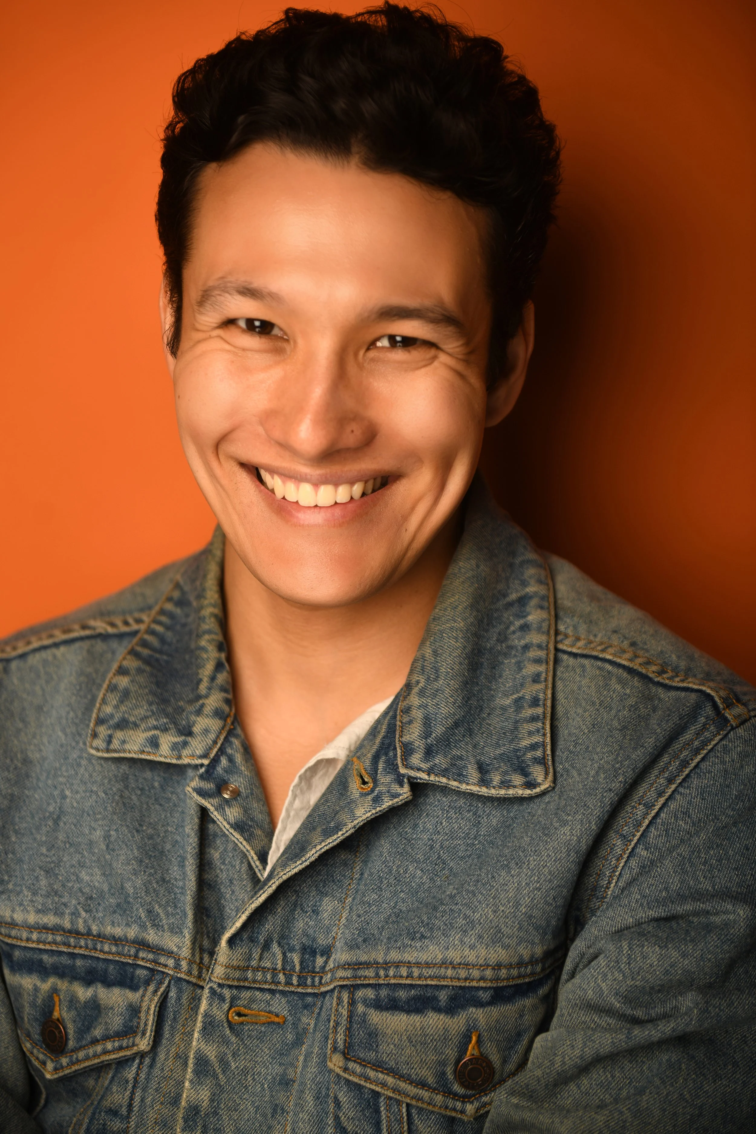 A young man smiling with a denim jacket against an orange background.