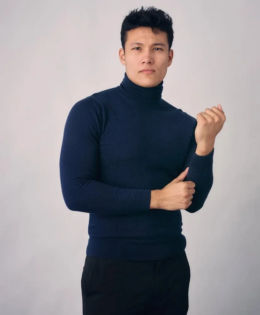 A young man with dark hair wearing a navy turtleneck sweater, looking serious, with his right fist clenched and left hand resting on his right arm against a neutral background.