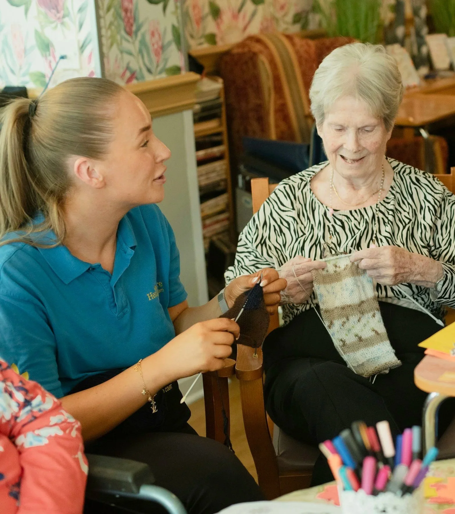 A young caregiver and an elderly woman sit at a table, knitting together and smiling, in a cozy room with patterned wallpaper and books in the background.