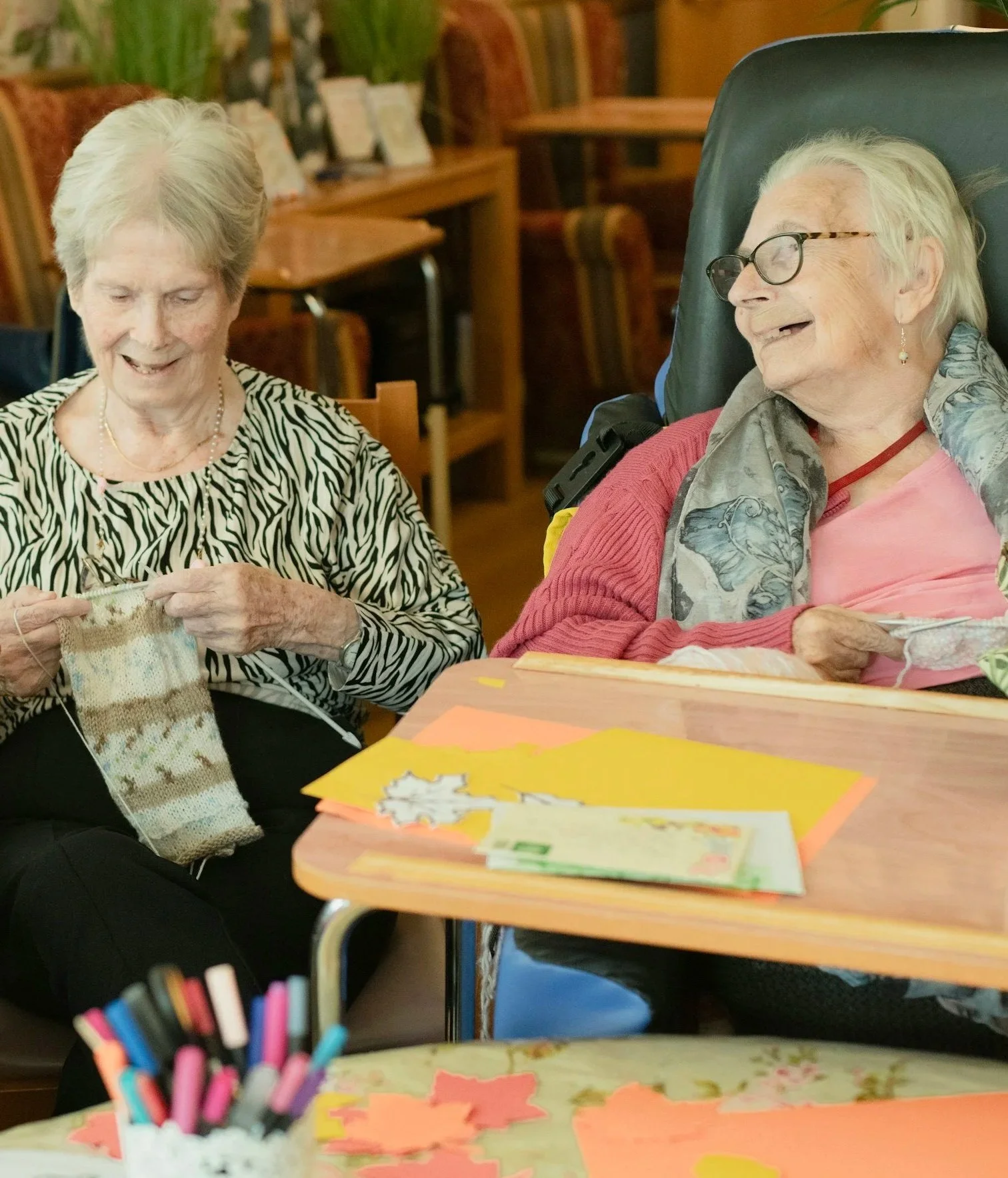 Two elderly women smiling and talking, one knitting and the other looking happy, sitting at a table with paper crafts and pens.