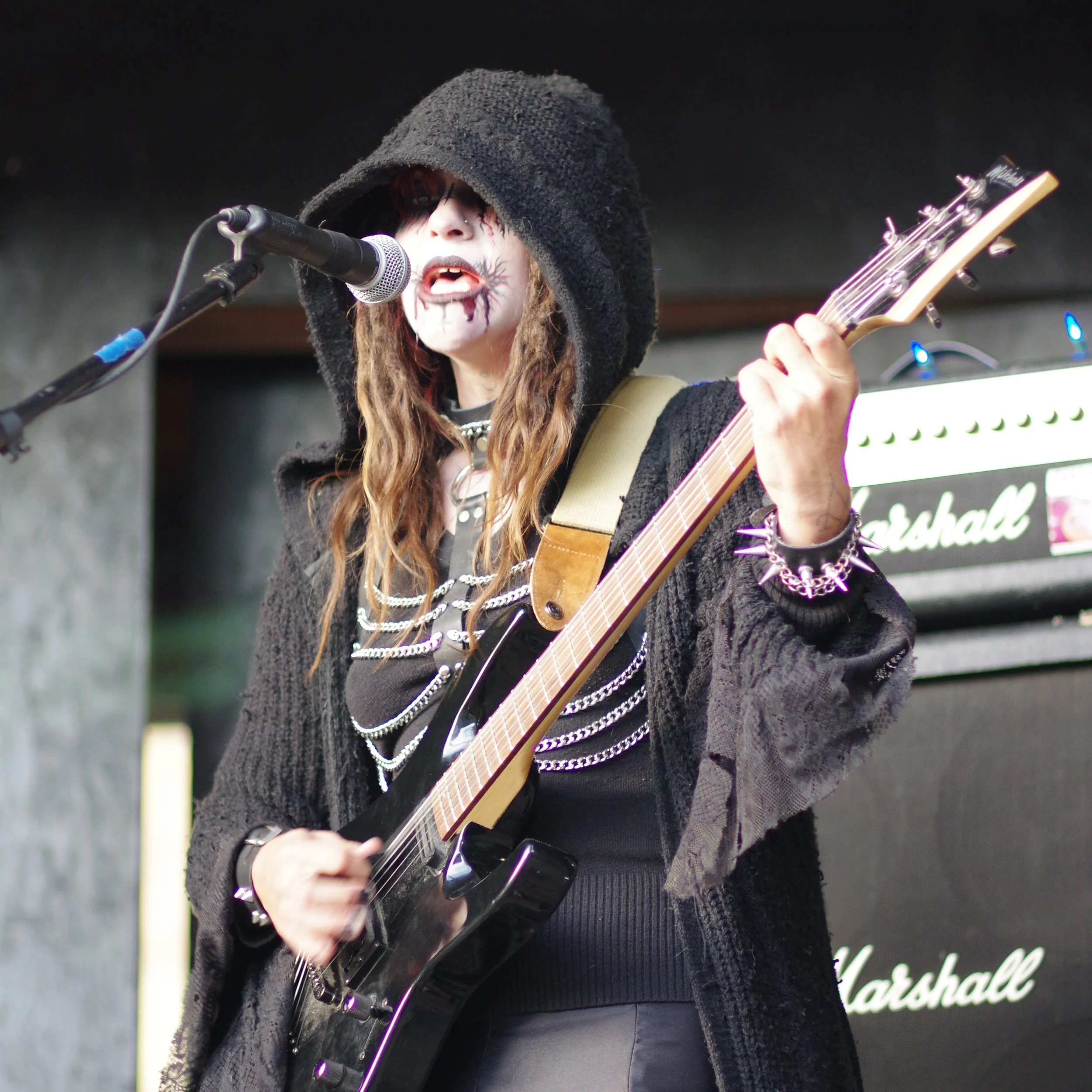 A metal musician named Xina Christel,  long hair wearing corps paint with a black hooded cloak, playing an electric guitar and singing into a microphone on stage, with a Marshall amplifier in the background in Pagosa Springs Colorado.