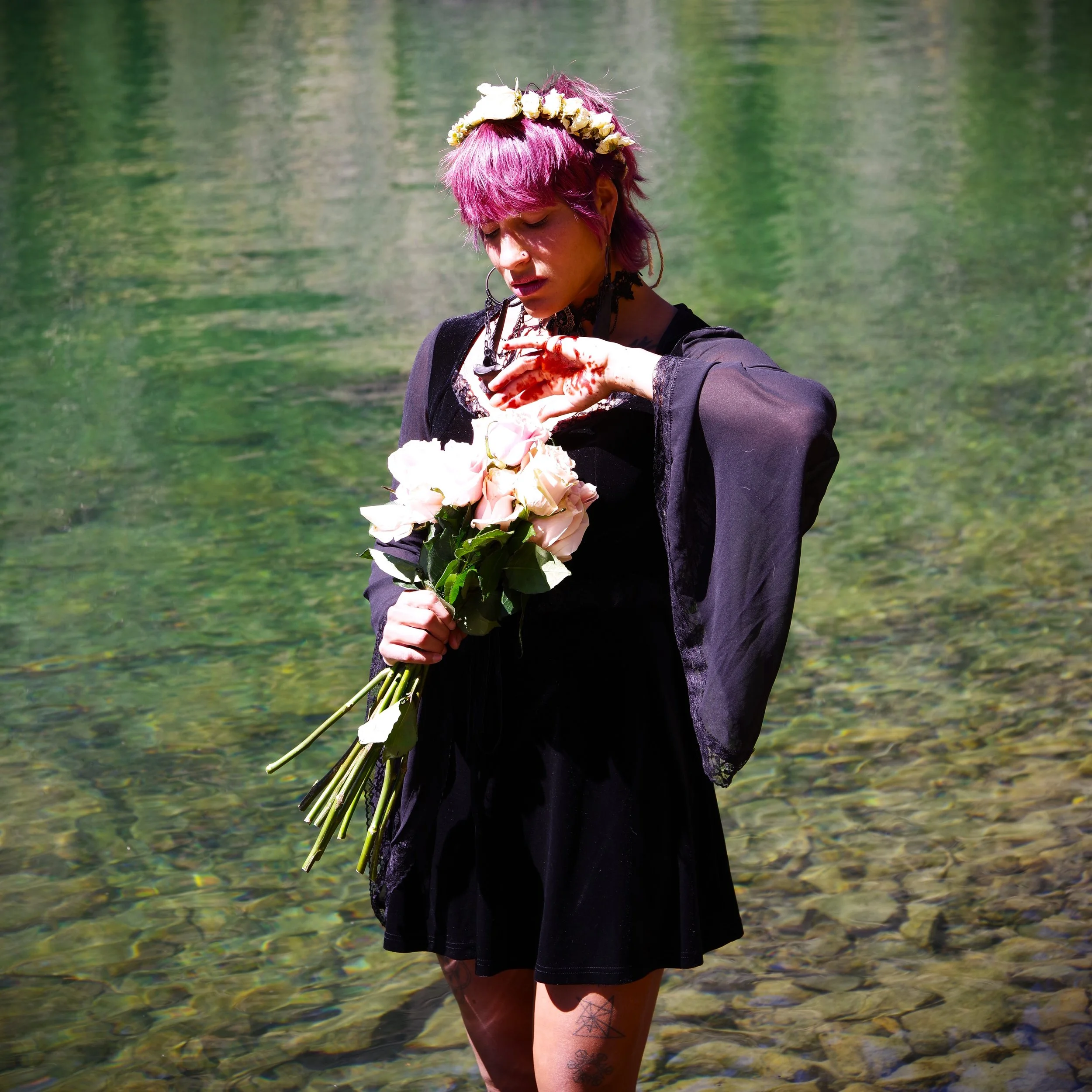 A woman named Xina Christel, wears a flower crown, pink hair and a gothic black dress, standing in a turquoise lake (Crater lake) in Colorado. She expresses pain and sorrow by holding a bouquet of roses with blood on her left hand.