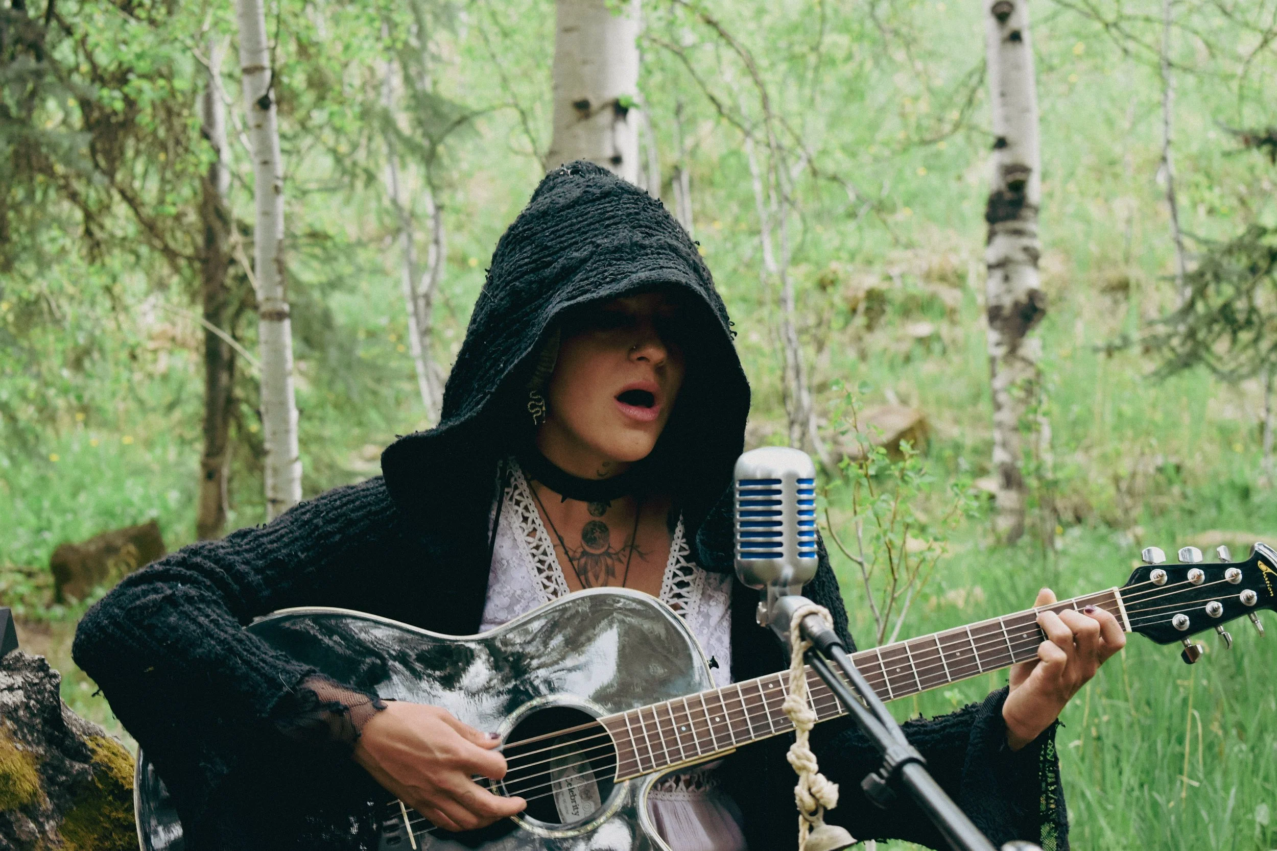 A musician named Xina Christel,  in a black hooded cloak plays an acoustic guitar and sings into a microphone in a forested area in Colorado.