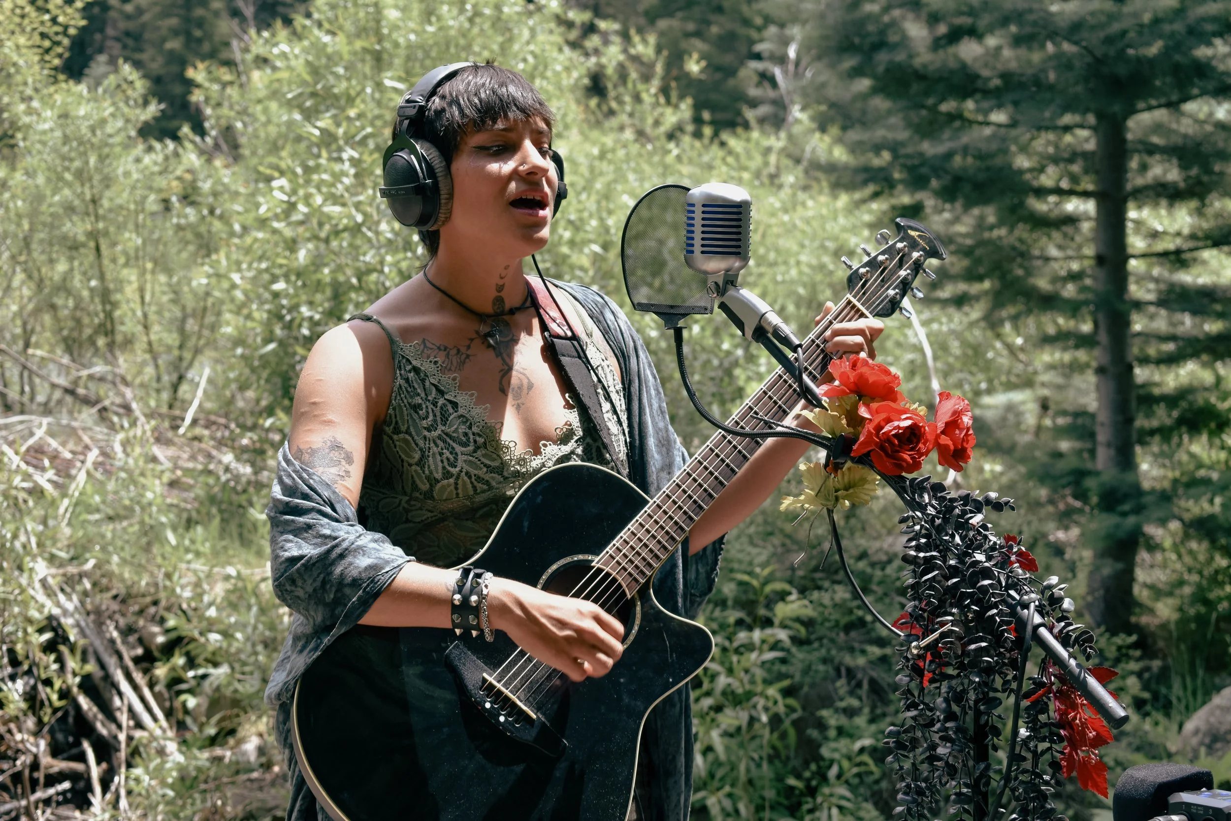 A woman named Xina Christel with tattoos and short brown hair, wearing headphones and a nymph like top, singing into a microphone while playing an acoustic guitar decorated with red and black flowers, outdoors in a forest setting in Colorado.