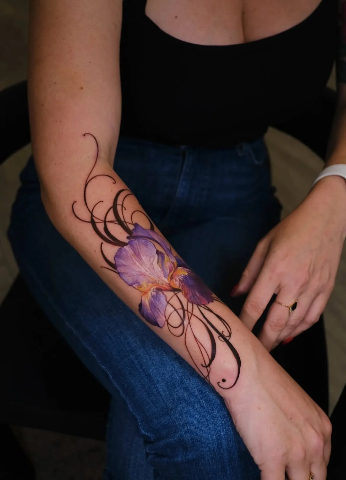 A tattoo of a purple iris flower with orange accents and black swirling decorative lines on a woman's forearm.