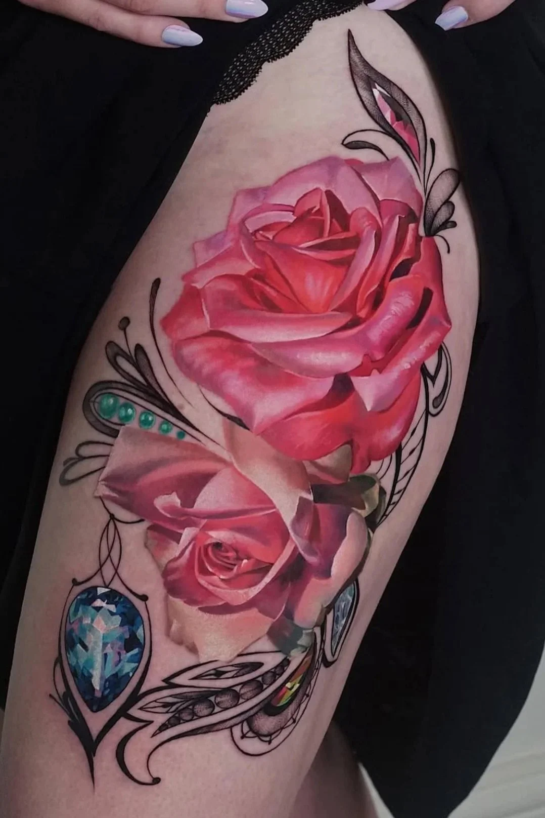 A colorful tattoo of pink and red roses with black outline accents, decorated with blue and green gemstones and abstract black line designs on a person's upper arm.