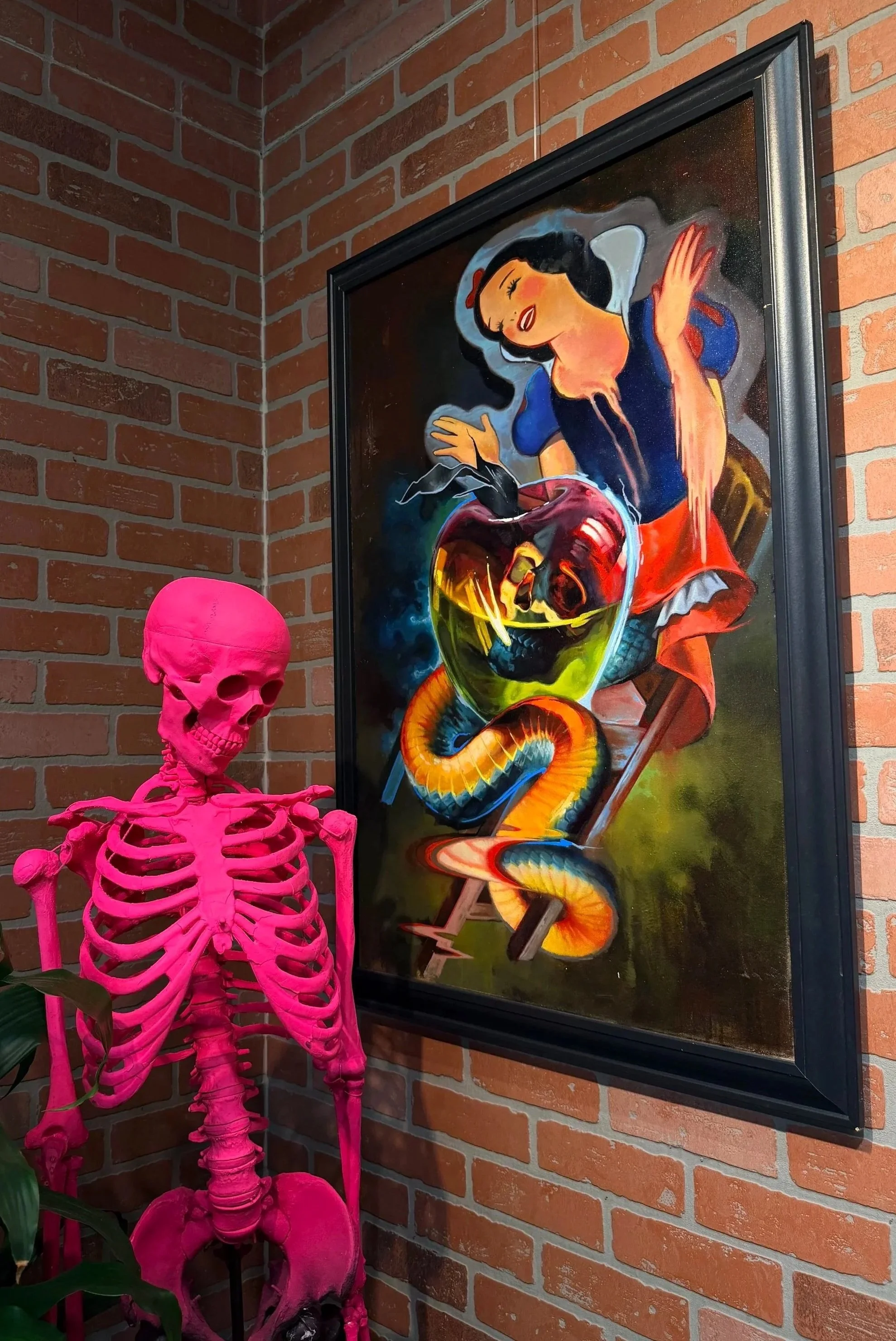 A pink skeleton statue standing next to a colorful, surreal painting on a brick wall. The painting depicts a woman with black hair and red lips, sitting on a chair with a snake wrapped around her, a jar with a heart inside, and vivid, dark background elements.