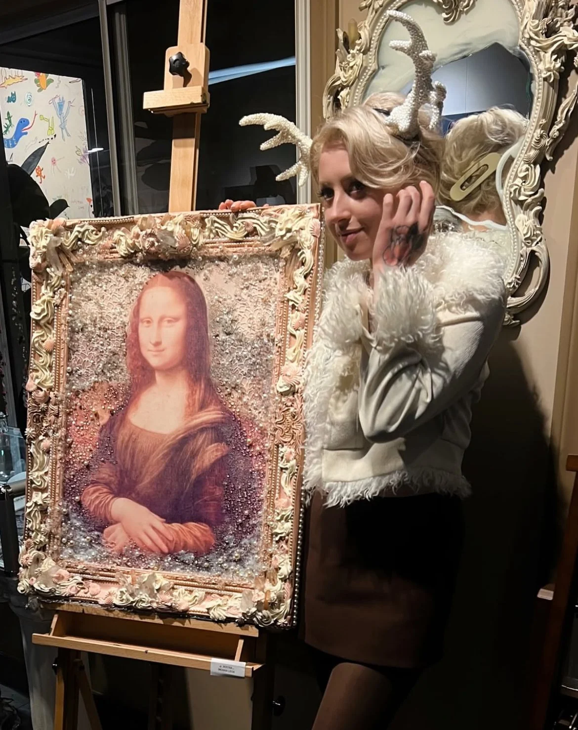 A woman wearing a white, fluffy jacket and antler headband with sparkles poses next to a decorated portrait of the Mona Lisa, with her hand resting on her face.