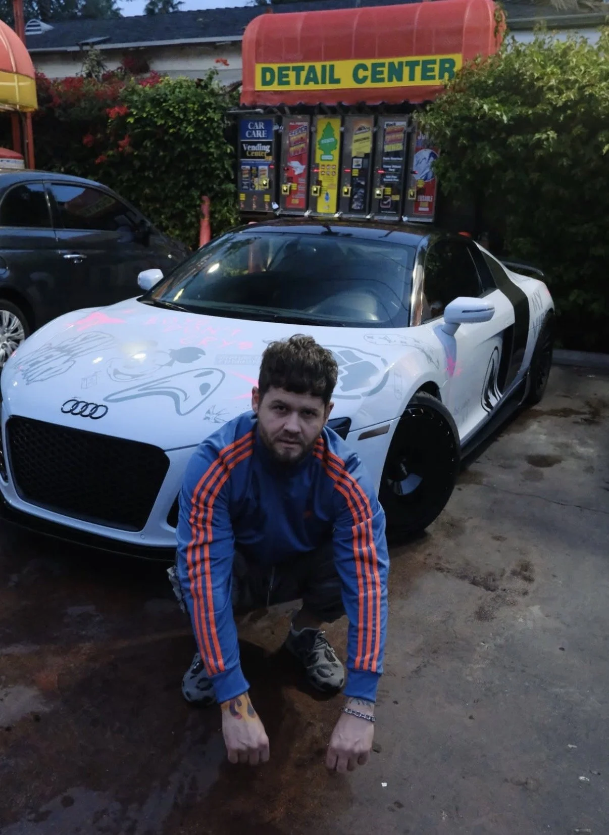 Man squatting in front of a white sports car with artwork on the hood at a car detail center, with vending machines in the background.