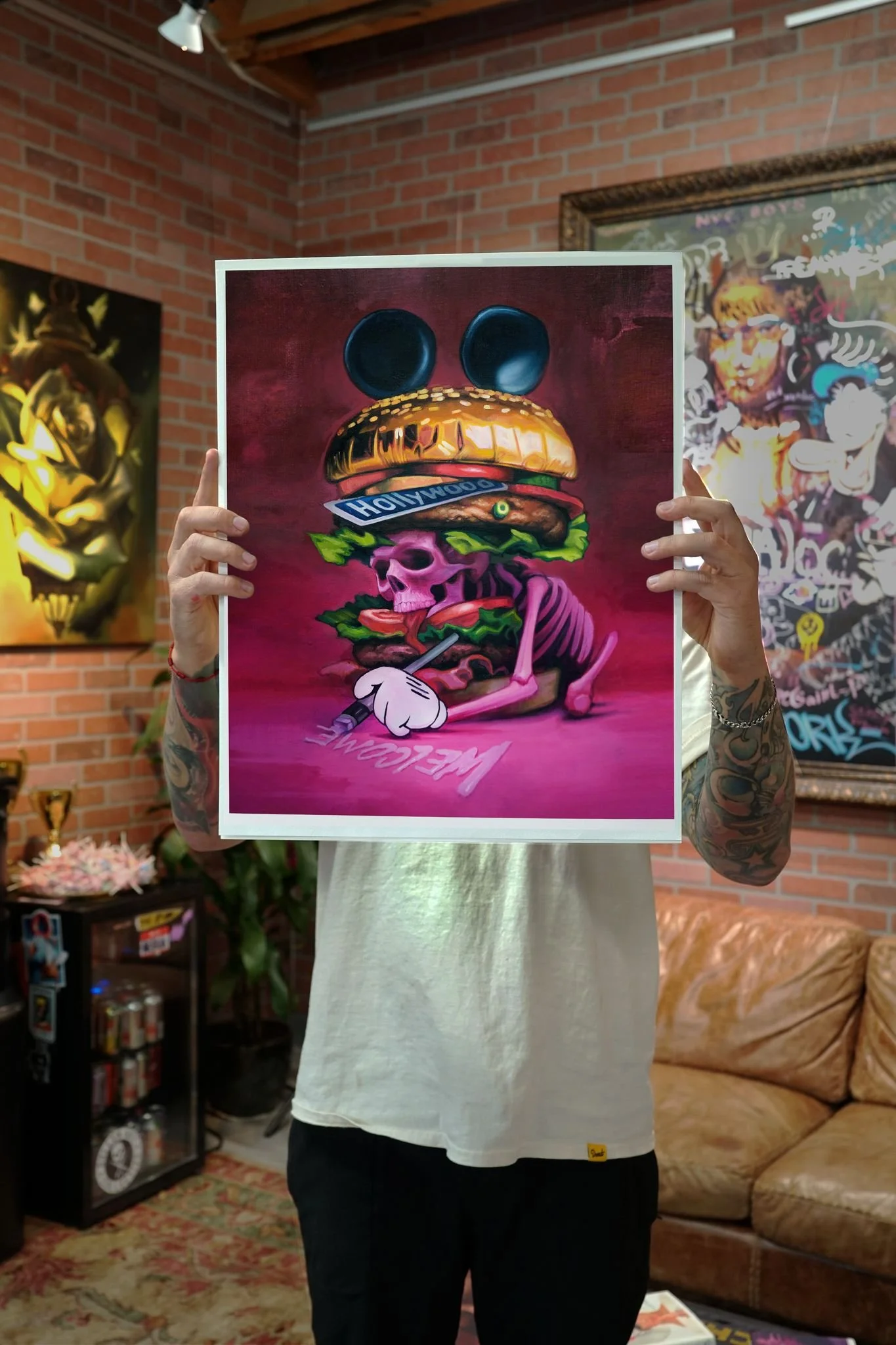 Person holding up colorful artwork featuring a stylized skull with a burger, Mickey Mouse ears, and the word 'Hollywood' on a banner, in an art gallery with brick walls and various artwork in the background.
