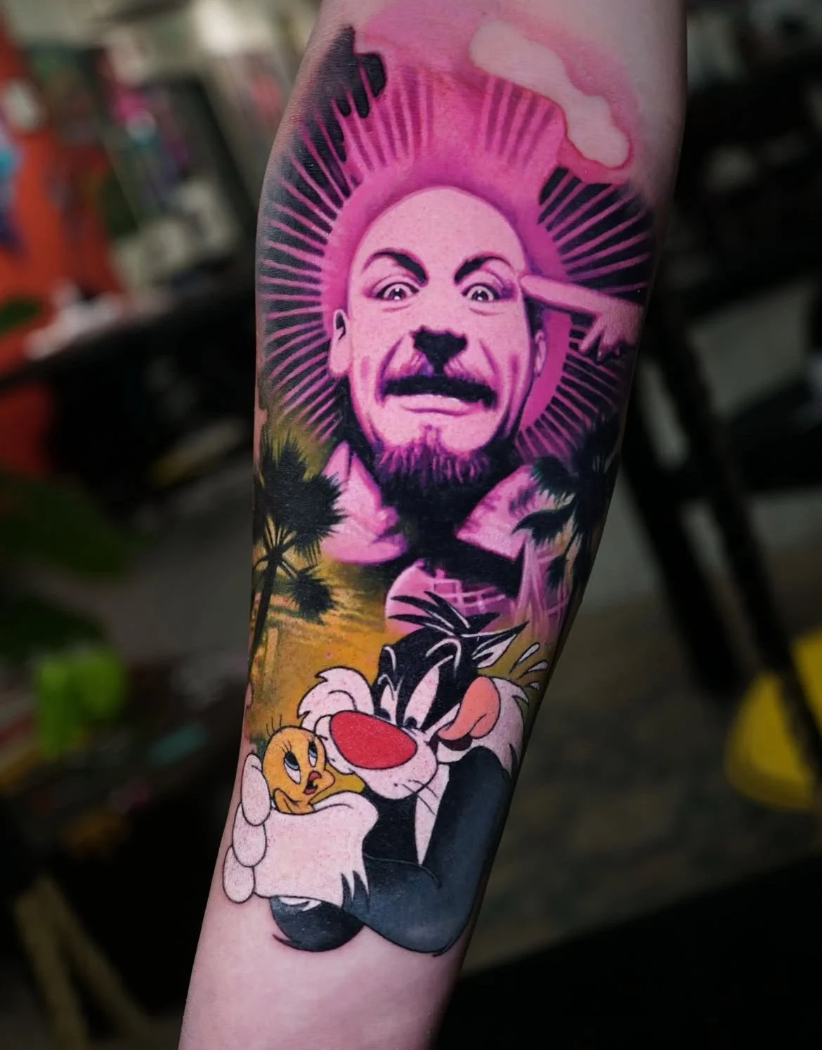 Tattoo of a man's smiling face with a pink background and rays, Tom and Jerry characters, Tom hugging Jerry, with palm trees and sunset in the background.