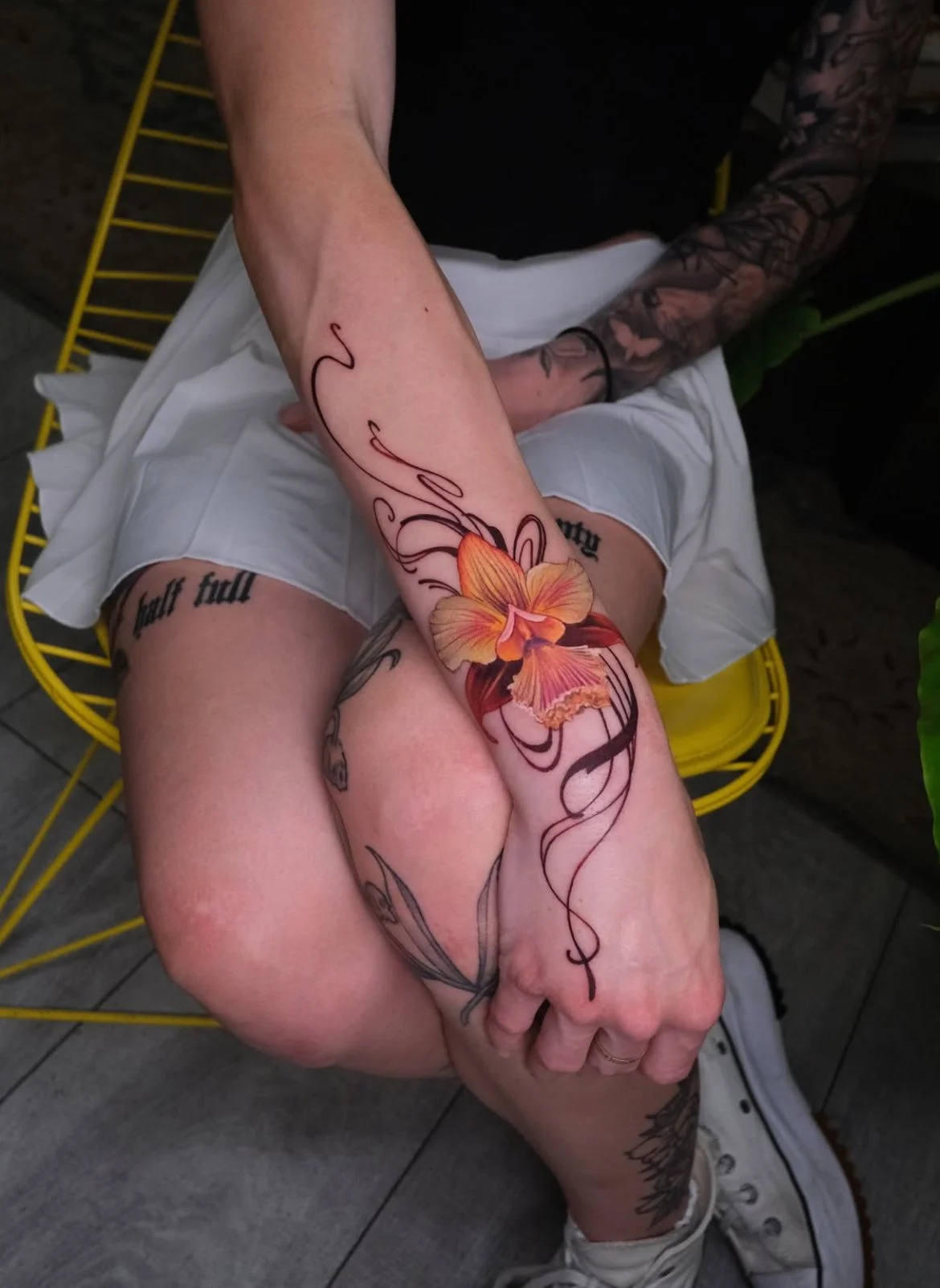 A person with colorful floral tattoos sitting on a yellow chair, wearing white shorts, and white sneakers.