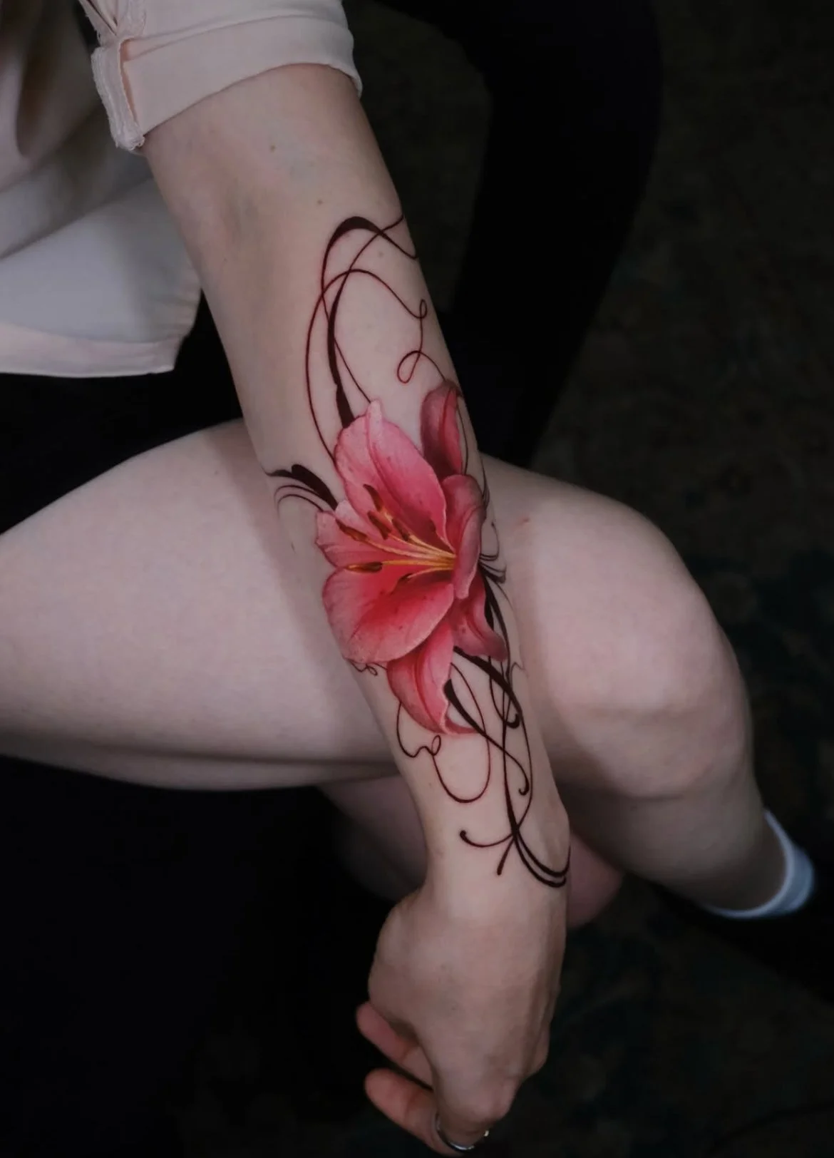 A person's arm with a detailed pink lily flower tattoo and black decorative swirls.