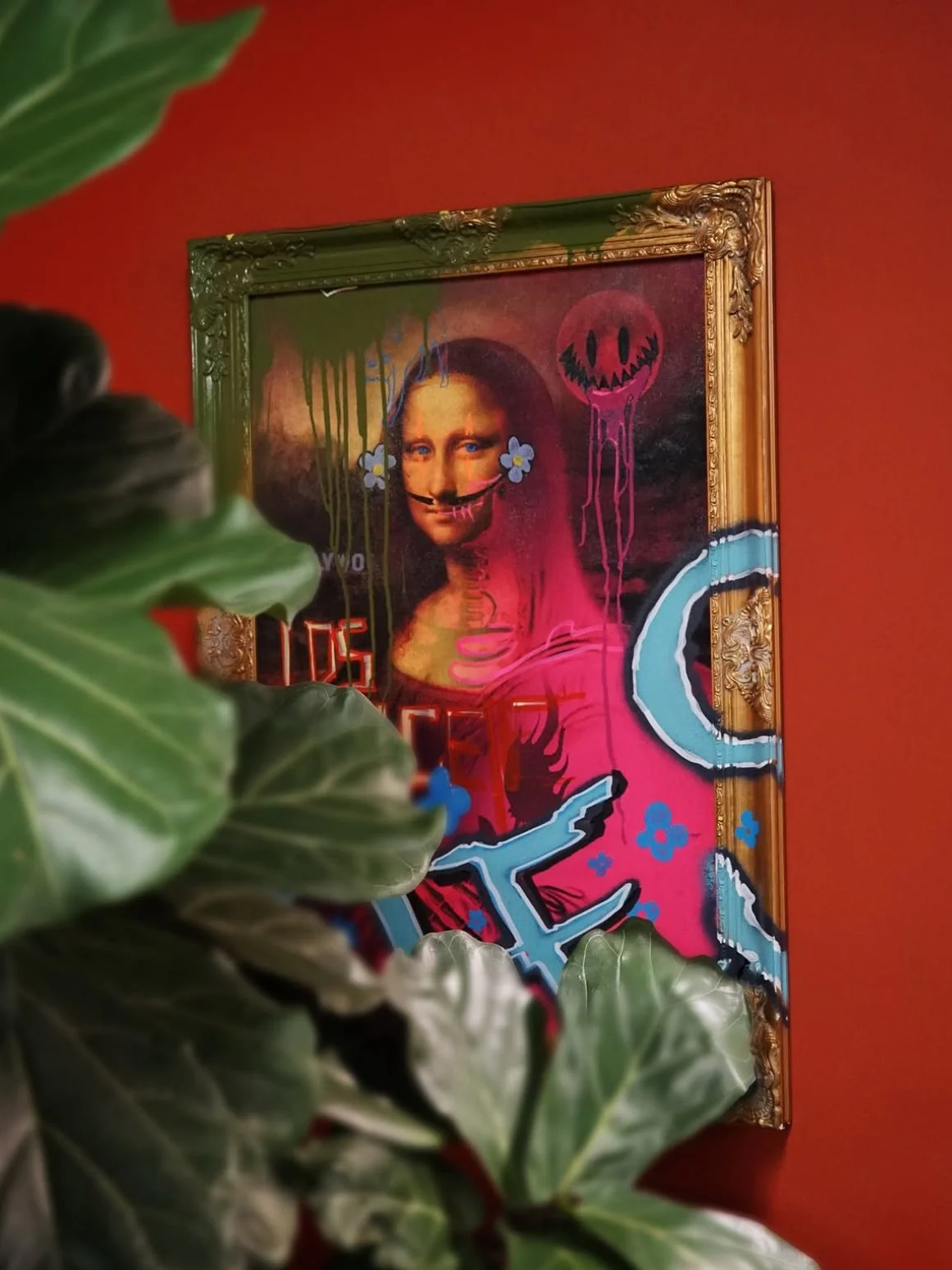 A framed painting of the Mona Lisa combined with colorful graffiti-style street art, including a dripping smiley face, blue flowers, and the word "LOVE," with green leaves in the foreground and a red wall in the background.