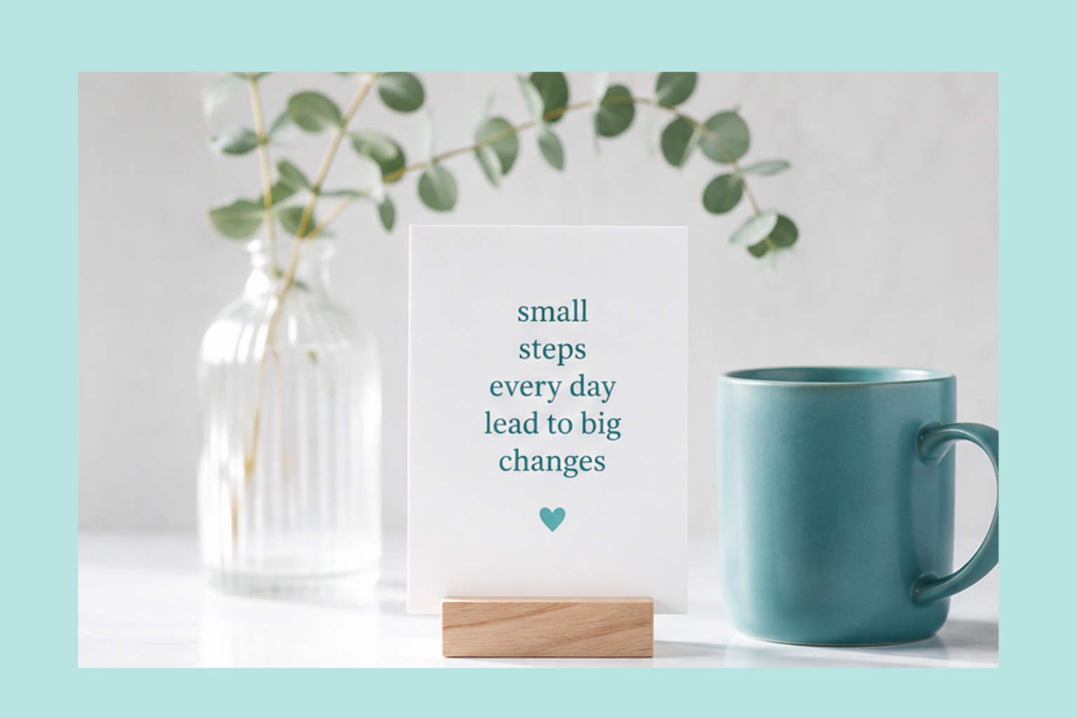 A white card with an inspirational quote on small daily steps in teal text, placed on a wooden stand, with a green vase holding eucalyptus branches in the background and a teal mug on the side.
