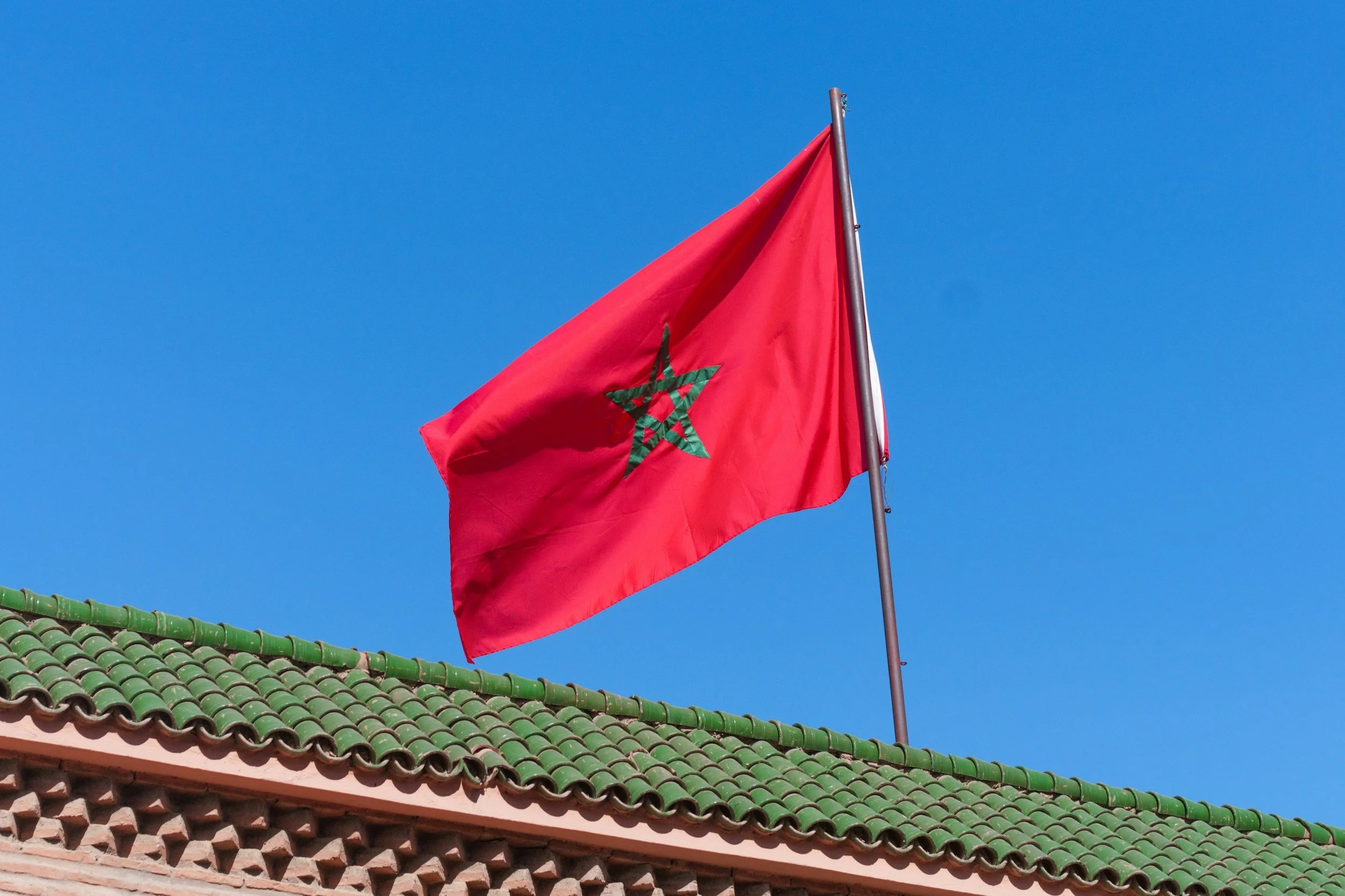 Drapeau du Maroc flottant au vent contre un ciel bleu, au sommet d'un bâtiment avec un toit en tuiles vertes.