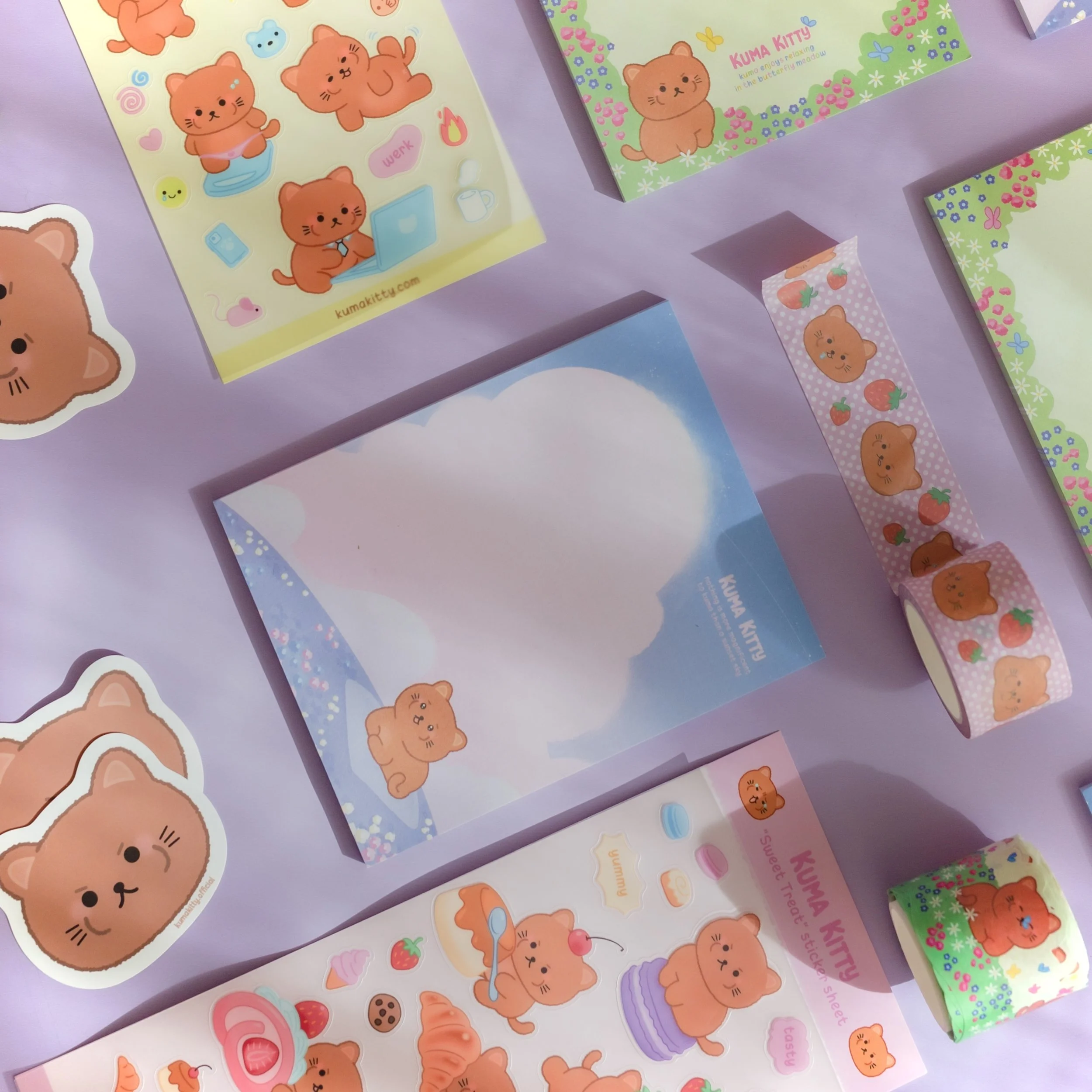 Various KUMA KITTY stationery items including stickers, washi tapes, and sheets, all themed with cute, orange bear and cat characters, displayed on a pastel purple background.