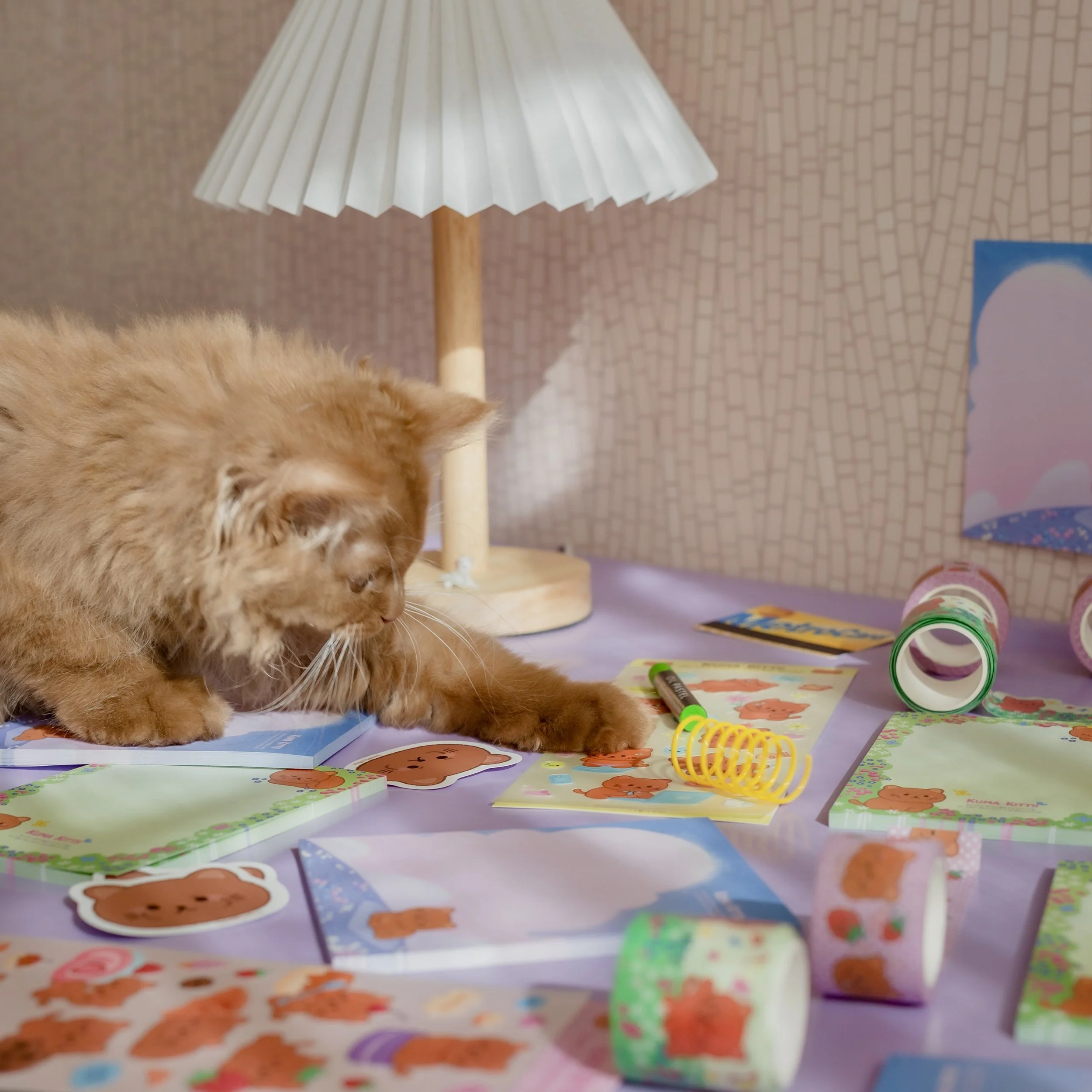 Orange cat surrounded by colorful tape, stickers, and drawing supplies on a table with a lamp in the background.