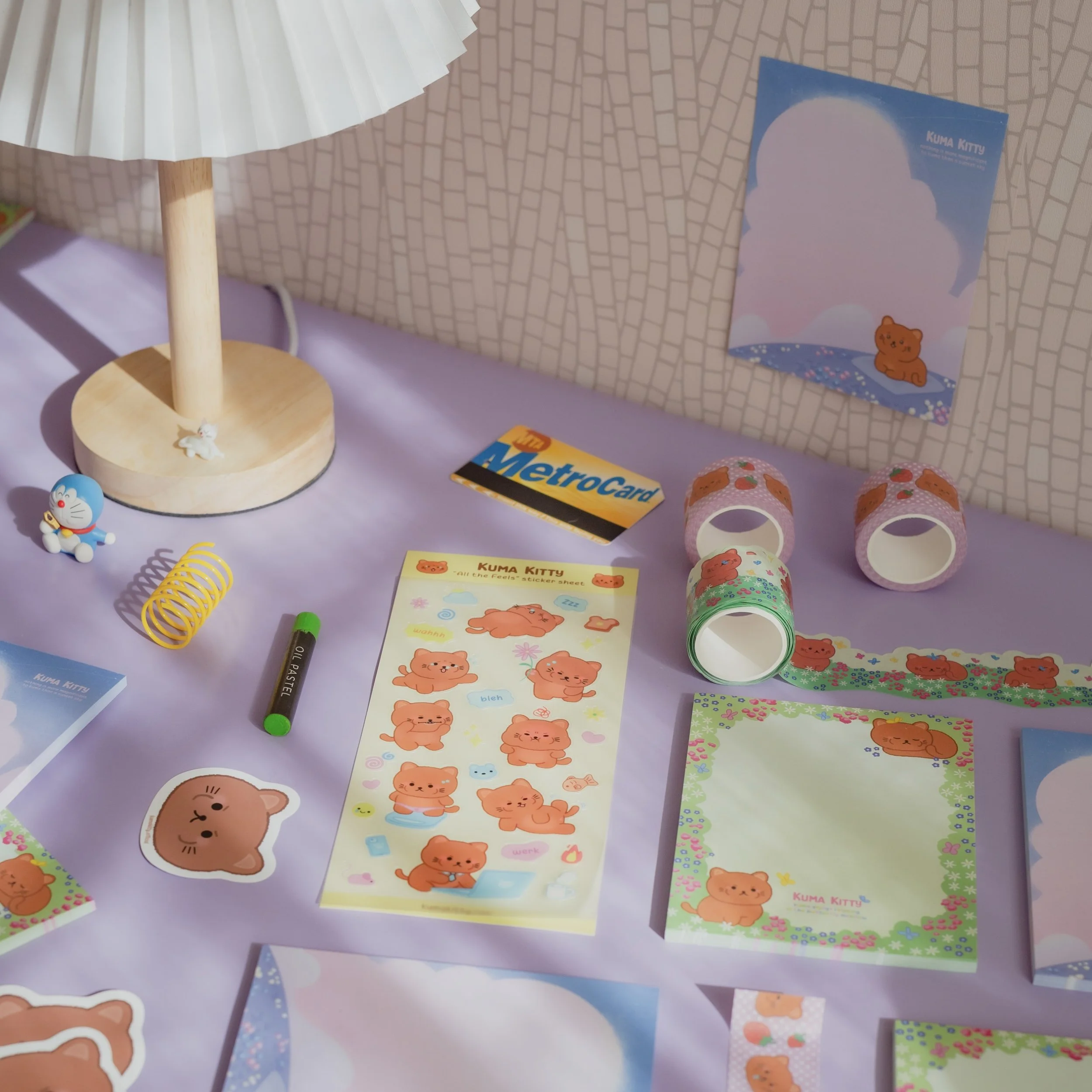 A collection of Kuma Kitty stationery and accessories on a lavender table, including a sticker sheet, notebooks, rolls of washi tape, a metrocards, a Doraemon figure, and a small lamp.