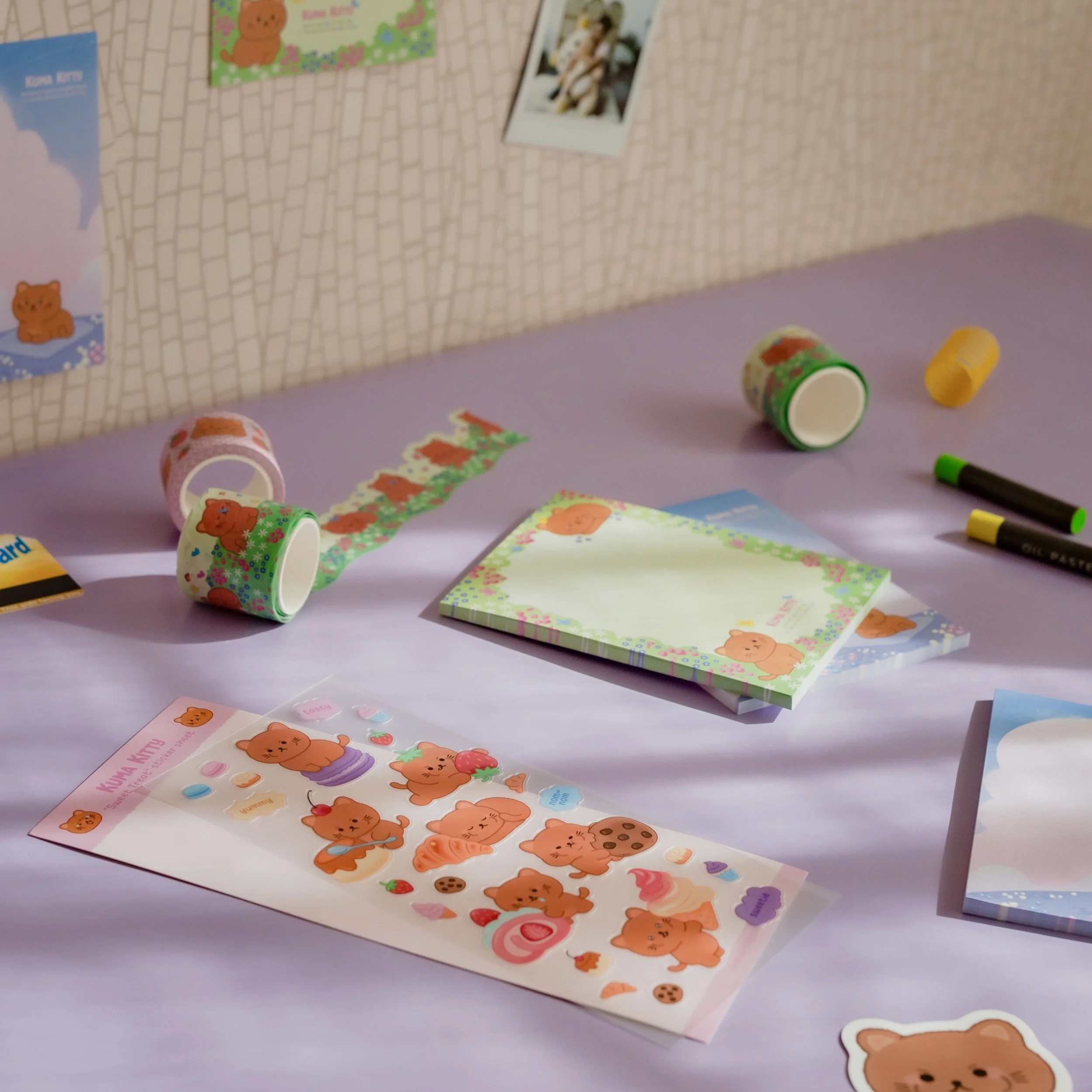 Decorative stationery with bear designs, stickers, notebooks, and markers on a purple table, with a wall and photos in the background.