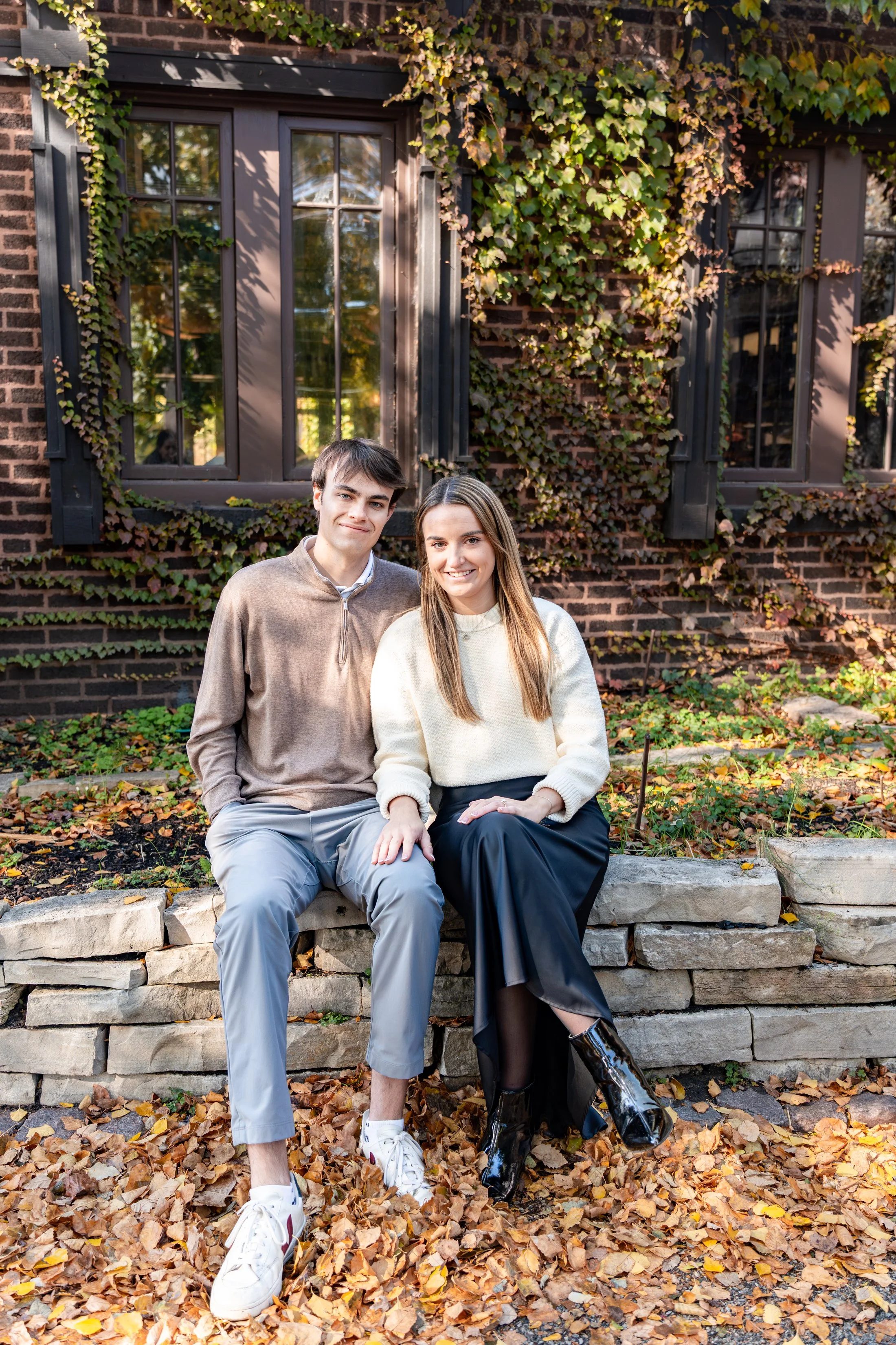 A young man and woman sitting on a stone wall outdoors with autumn leaves on the ground and a brick house with ivy-covered windows in the background.