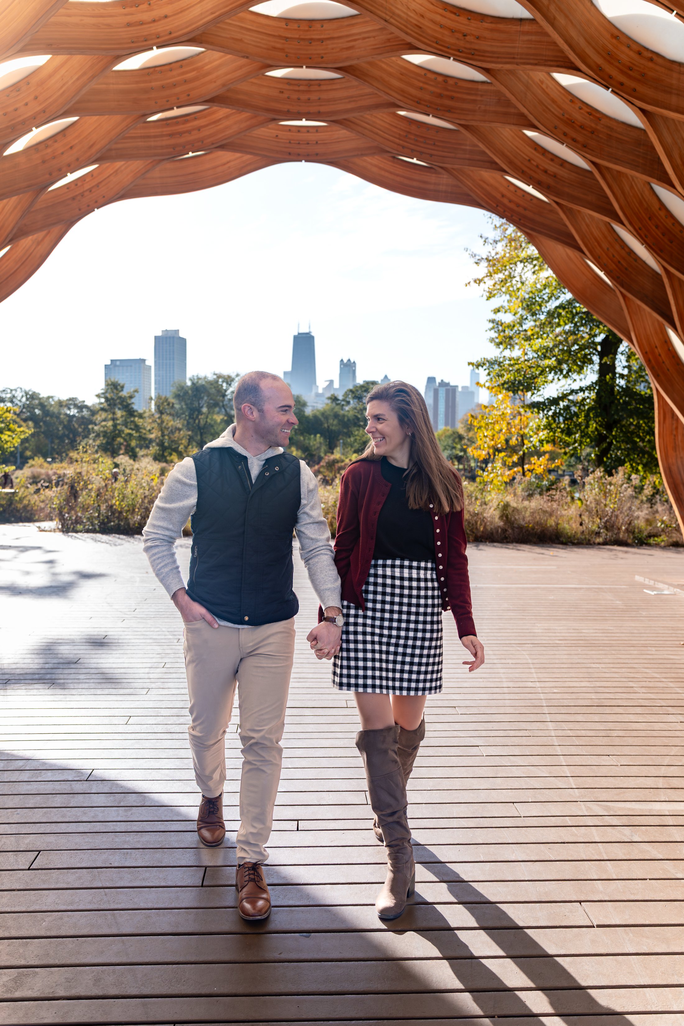 A couple walking hand in hand outdoors underneath a wooden archway, smiling at each other with a city skyline in the background during daytime.