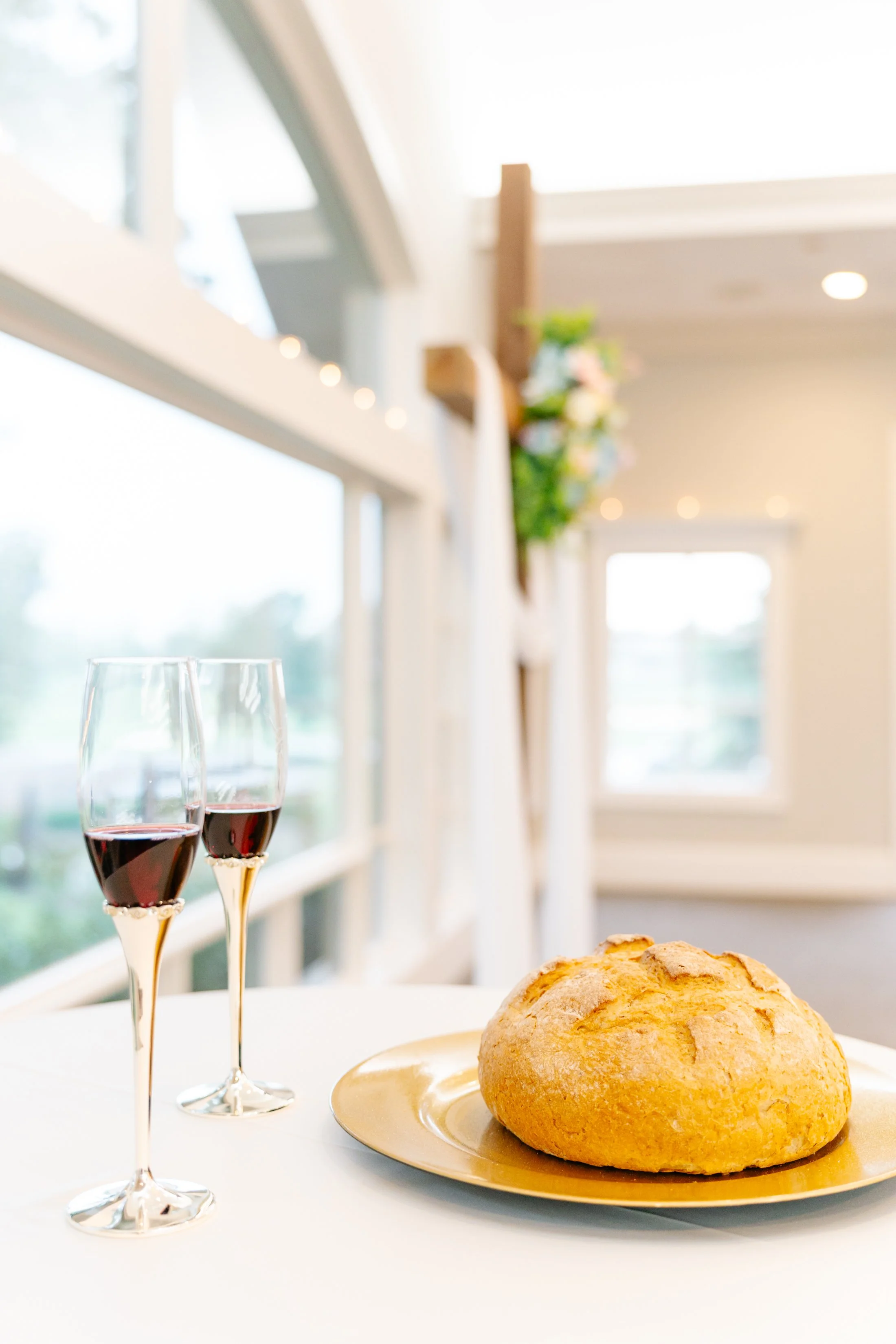 Two glasses of red wine and a loaf of bread on a white table near a window in a bright room.