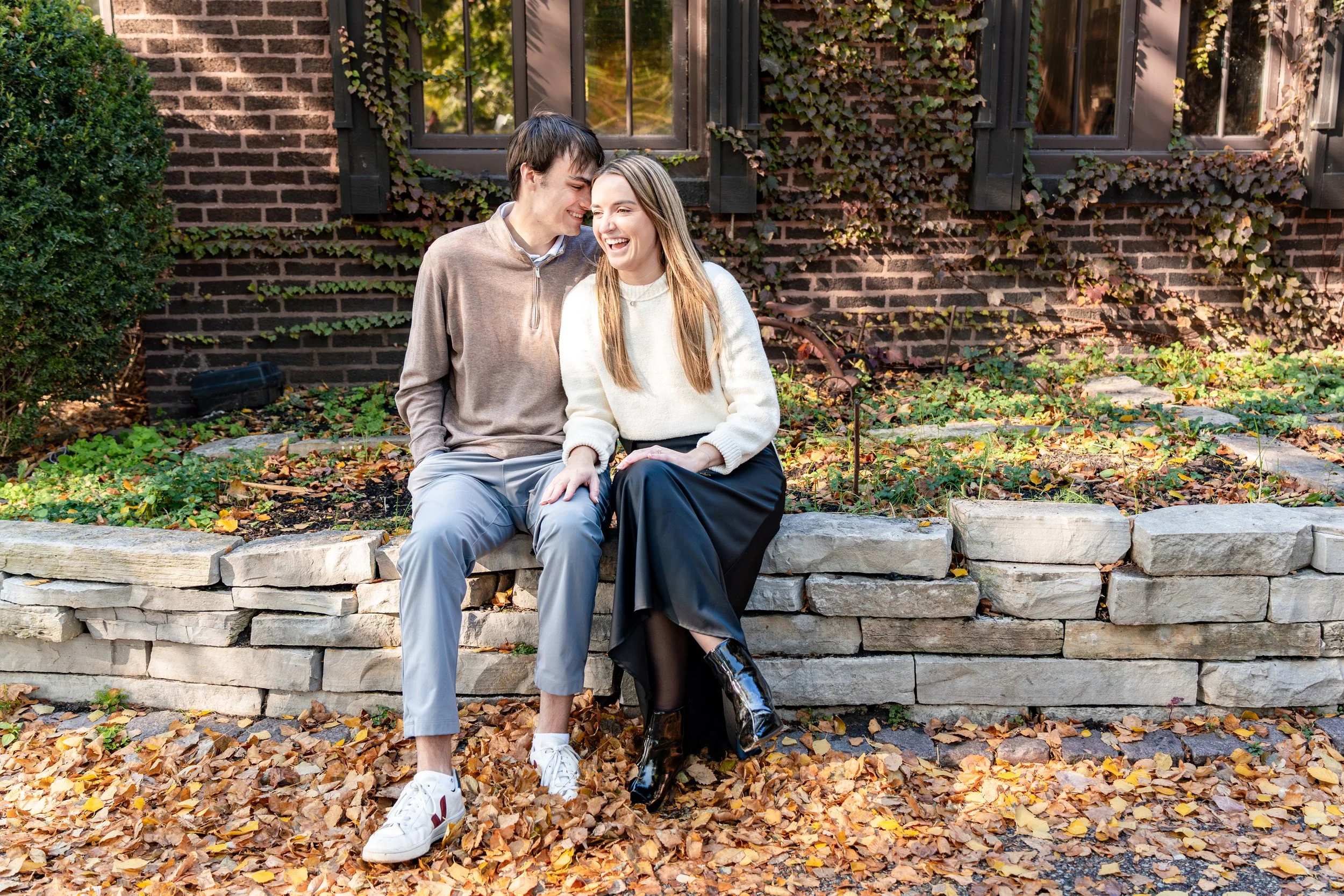 A young man and woman sitting together on a low stone wall outside, smiling and laughing, surrounded by fallen autumn leaves in front of a brick house with ivy.