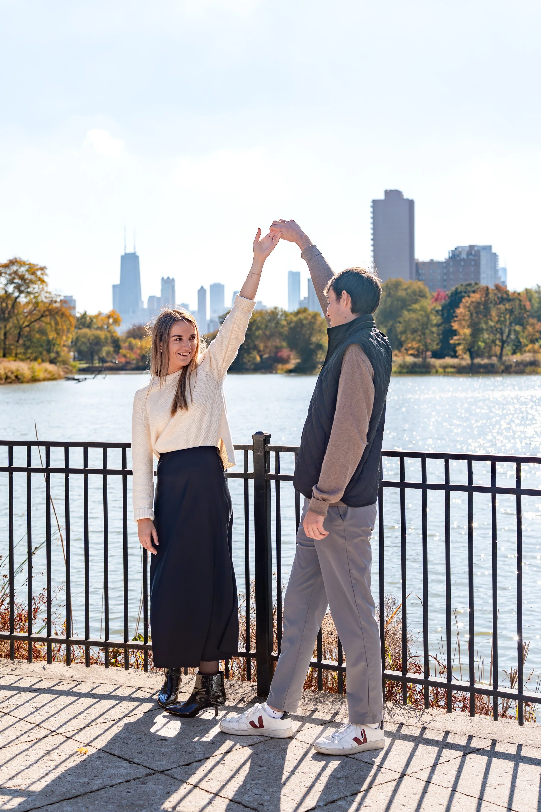 A young woman and a young man dancing by a river during autumn. The woman is smiling and wearing a cream sweater, a long black skirt, and black boots. The man is dressed in a grey and brown jacket, grey pants, and white sneakers. They are holding han