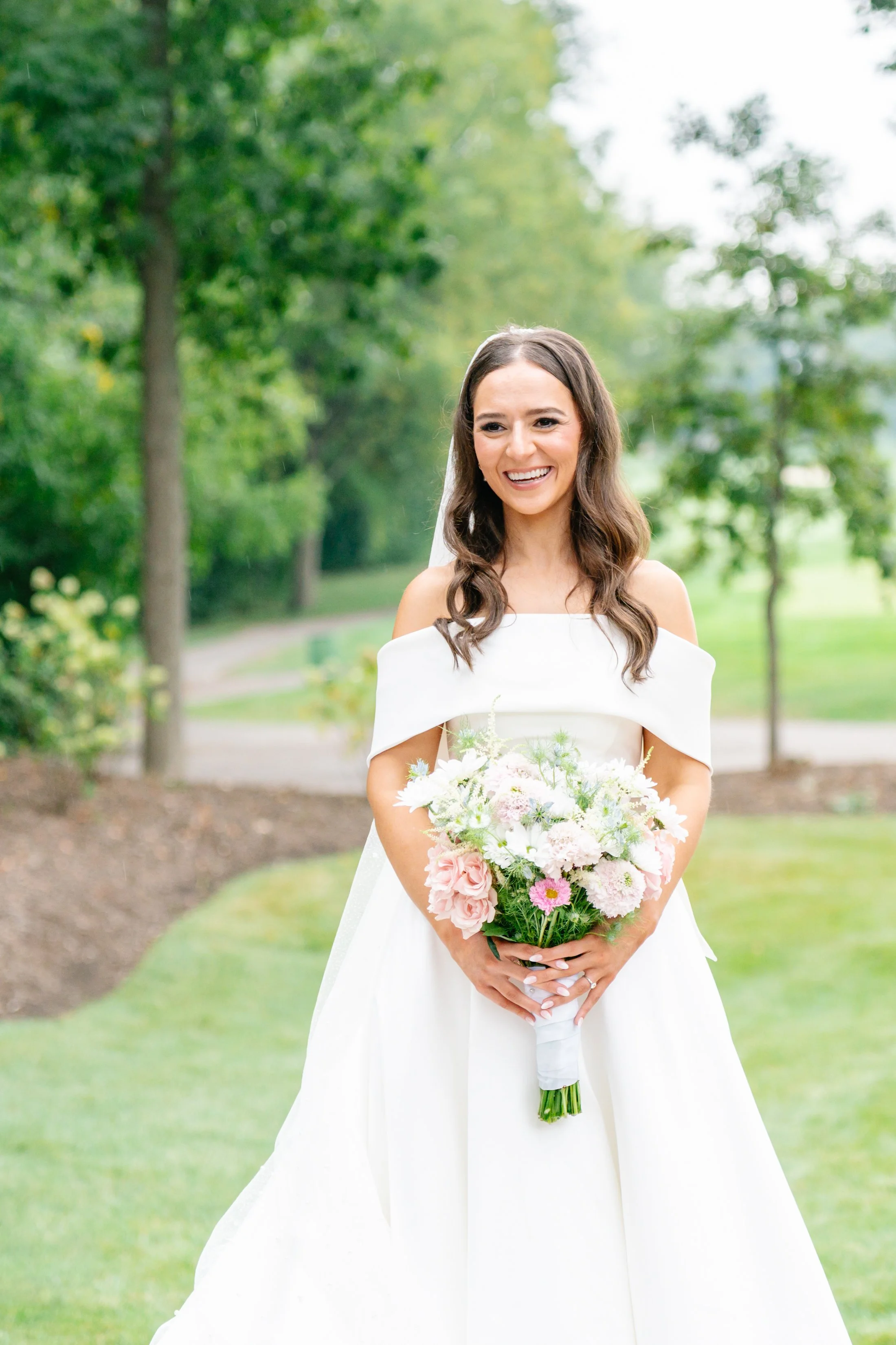 A smiling bride in a white wedding dress holding a bouquet of pink and white flowers outdoors on a grassy area with trees in the background.
