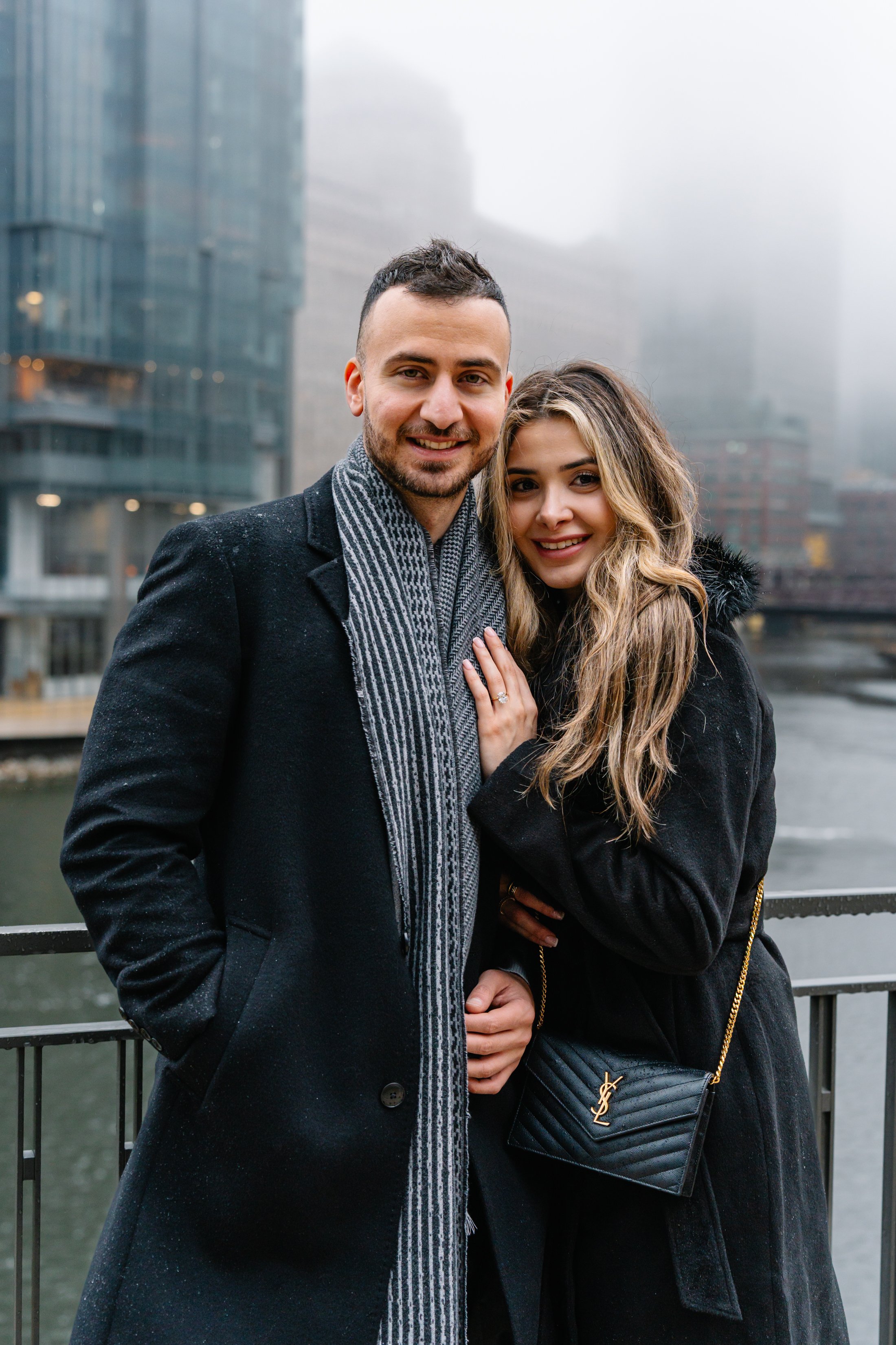 A smiling couple standing close together outdoors in a city with modern glass buildings, wearing winter coats on a rainy day.