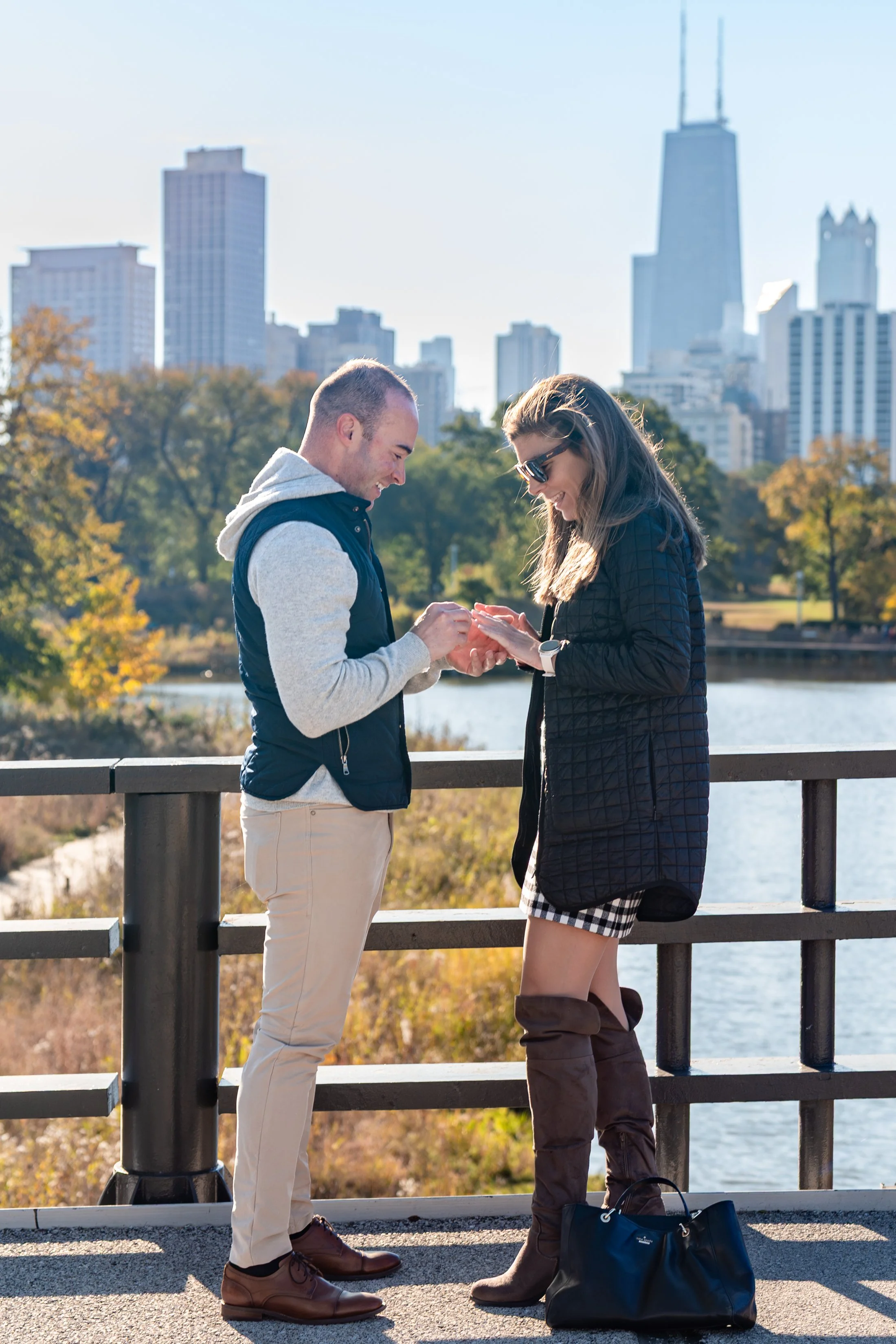 A man and a woman stand by a river, exchanging engagement rings with a city skyline in the background.