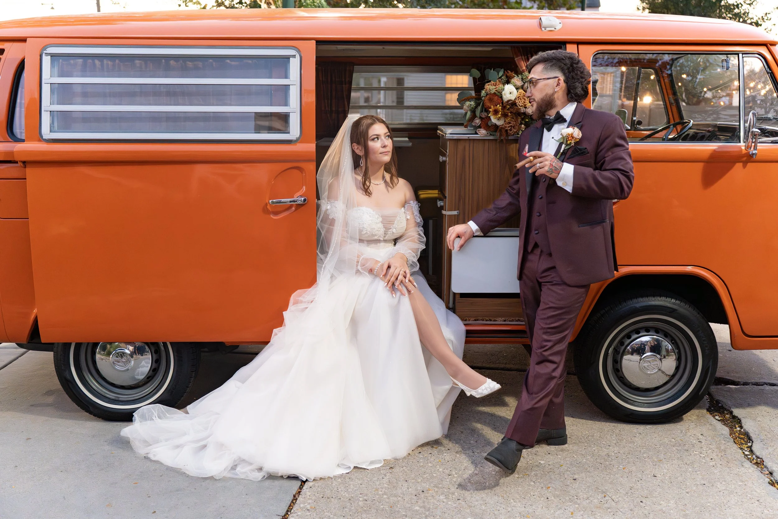 A bride in a white wedding dress sitting on the step of an orange vintage van, looking at a groom in a maroon suit with a bowtie, holding a cigar, standing next to her. The van has a wooden interior with flowers, and the scene takes place outdoors du
