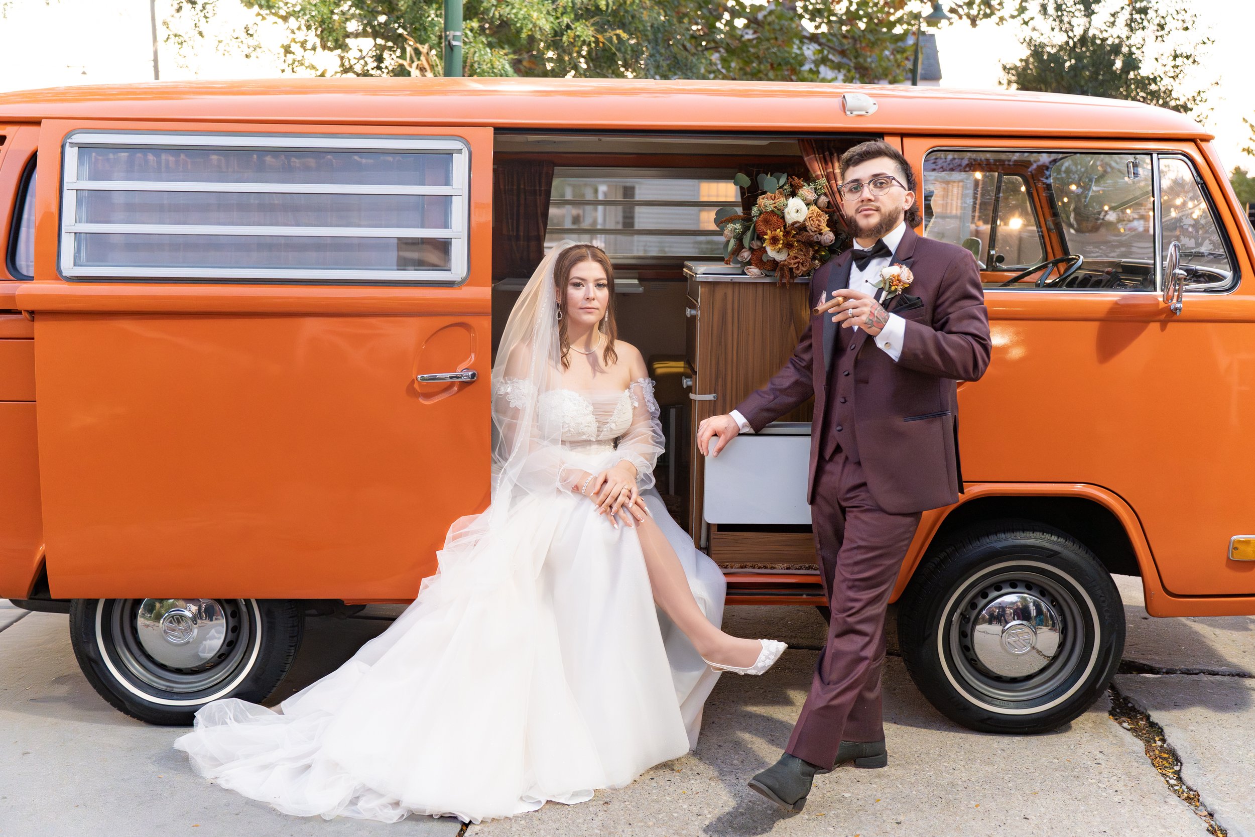 A bride in a white wedding dress and veil sitting on the step of an orange vintage van, with a groom in a burgundy suit and bow tie standing beside her, smoking a cigar and holding a glass, with a bouquet and decorative items inside the van.