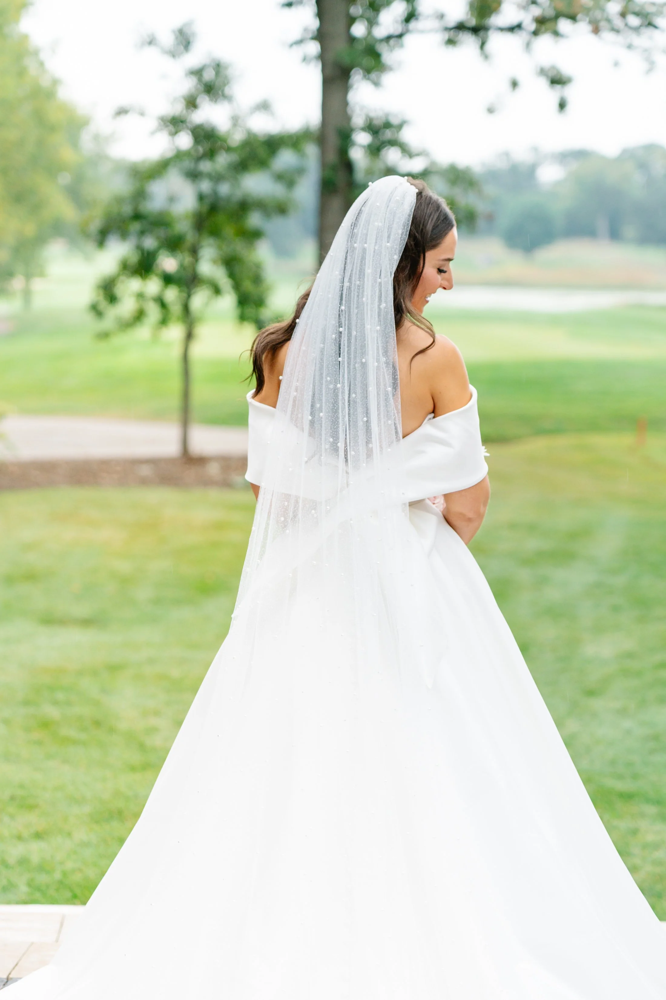 A bride in a white wedding gown with a long veil, standing outdoors on a grassy area, smiling and gently holding her off-shoulder dress, with trees and a body of water in the background.