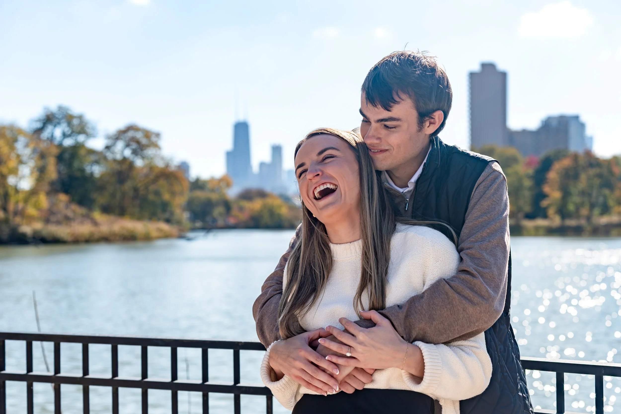A young couple hugging and laughing near a body of water with city buildings in the background.